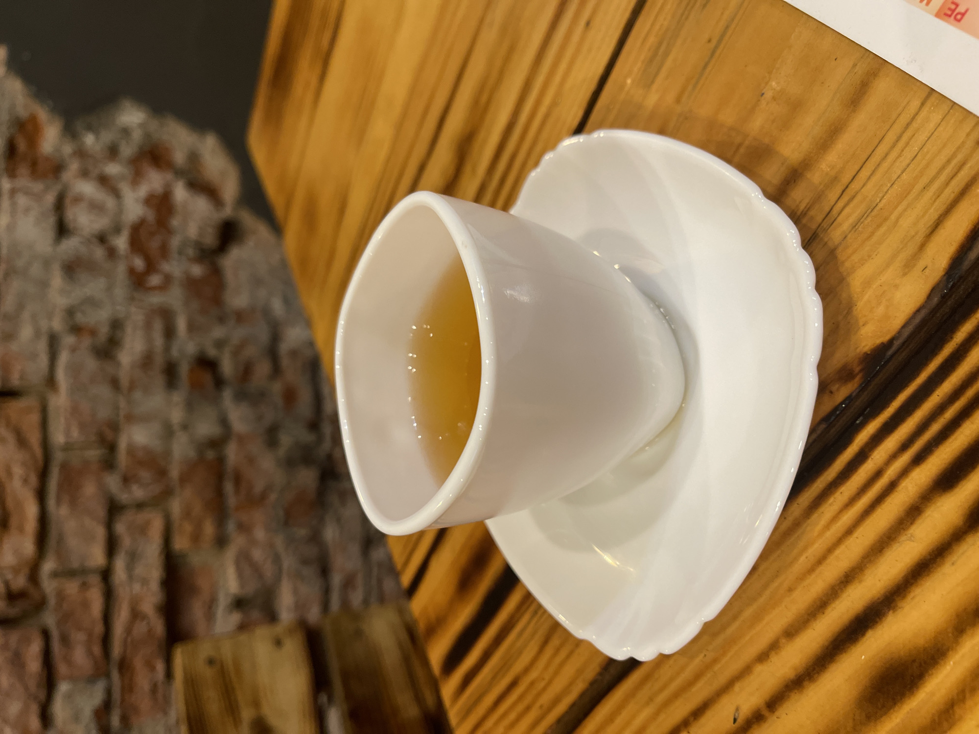 This scene, located in the city of Kaliningrad, Russia, features a single white ceramic teacup and saucer positioned on a wooden table. The teacup holds an amber-colored liquid, likely tea, with minor light reflections visible on its surface. The table exhibits distinct, roughly parallel dark grain patterns against a warm brown-orange wood. The background consists of an out-of-focus brick wall, showing reddish-brown bricks with lighter mortar. A darker, indeterminate area is visible in the upper right background, while a blurred wooden object, possibly furniture, is present on the left. A small segment of a white paper document with red text, partially displaying "PE," is visible in the bottom right corner. No individuals are present in the image.