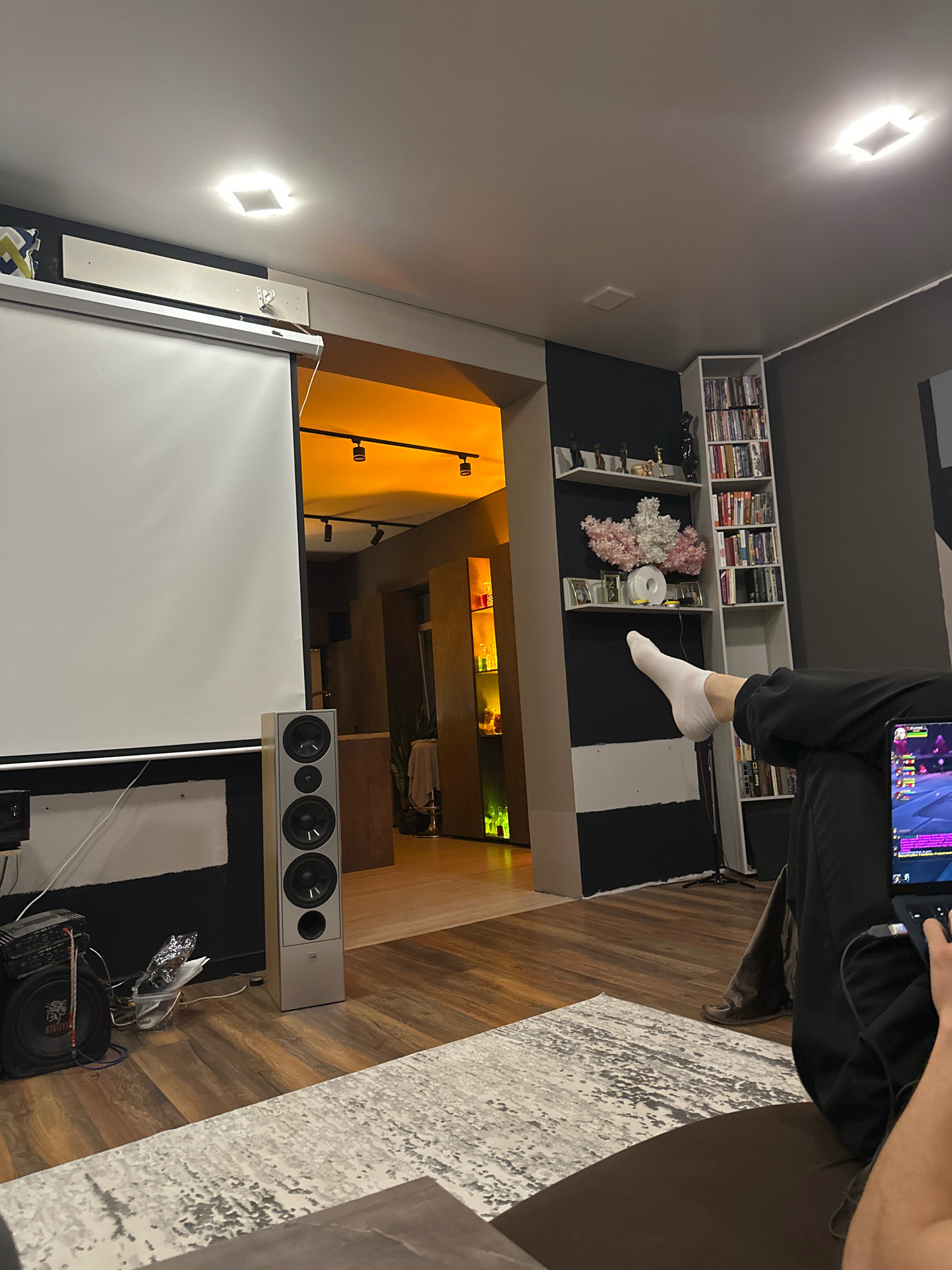 The image depicts an indoor domestic scene, likely a living room or home theater. A projection screen dominates the left side of the frame, with a tall, silver speaker positioned to its right. In the background, an open doorway reveals a hallway or adjacent room illuminated by warm-toned lights, with shelves displaying various items, including what appear to be bottles. To the right of the doorway is a tall, white bookshelf filled with books and decorative items. In the foreground, a person's legs, clad in black pants and white socks, are visible as they recline. The person is holding and looking at a laptop, suggesting they are engaged in an activity such as gaming or browsing. The floor is covered in wooden planks, with a patterned rug in the lower section of the image. The lighting suggests it is evening or nighttime. There is no discernible information within the image that places the scene specifically in Kursk, Russia.