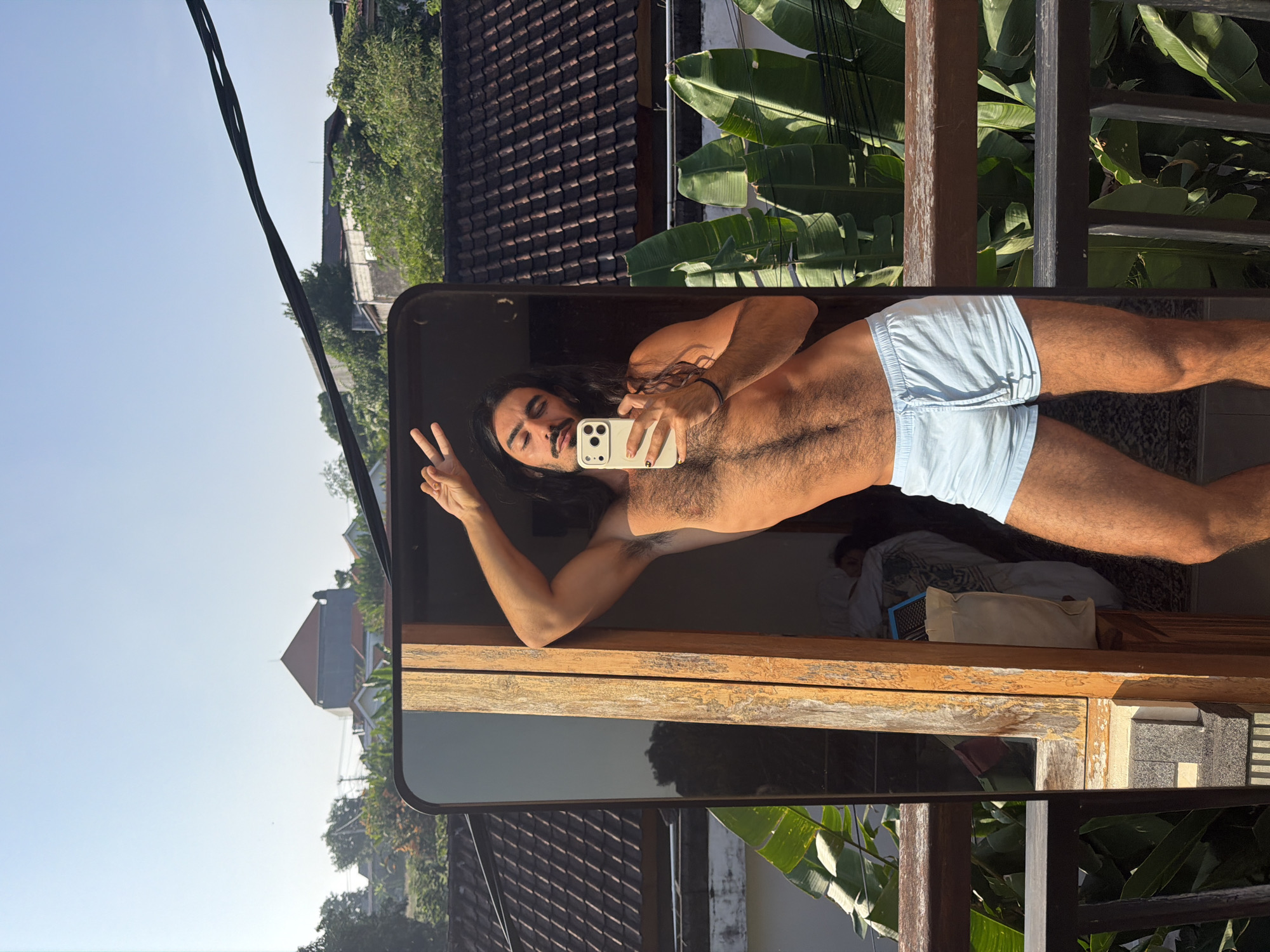 A man with long, dark hair and a beard stands on a balcony in North Kuta, Indonesia, taking a selfie in a mirror. He is shirtless and wearing light blue shorts. The man is leaning against a wooden frame, with his right arm raised and his left hand holding a smartphone. He is making a peace sign with his fingers. In the background, there are houses with tiled roofs, lush green foliage, and power lines. A person is visible sleeping in a bed inside the room behind the man. The lighting suggests it is daytime, likely morning or afternoon, due to the bright sunlight.