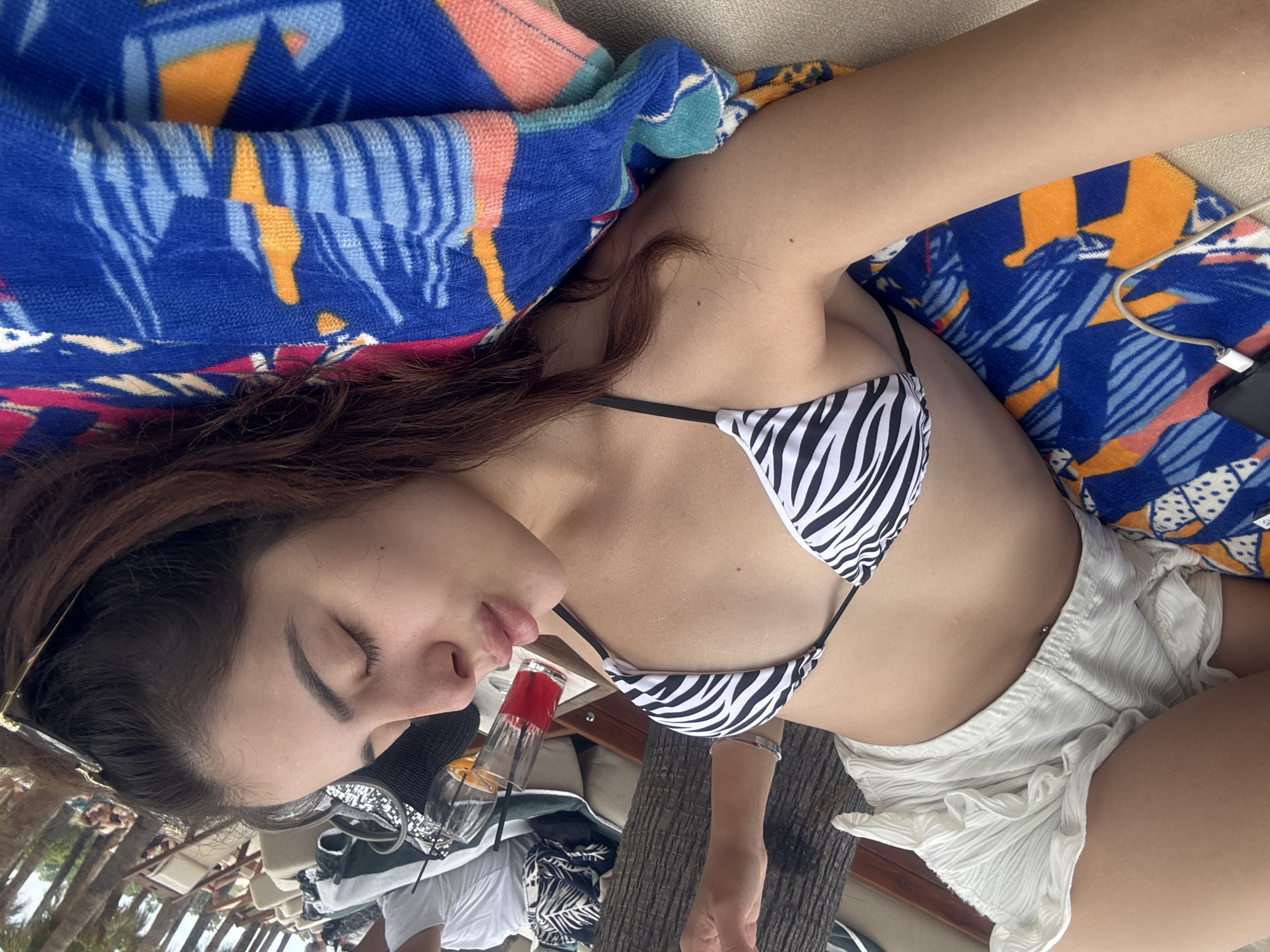 A young woman is lying on a beach towel, eyes closed, appearing to be asleep or resting. She is wearing a black and white zebra-print bikini top and white ruffled shorts. Her belly button piercing is visible. She is positioned on a lounge chair, and the background shows a beach setting with palm trees, other lounge chairs, and people in the distance, suggesting a resort or a public beach area. The setting is consistent with a location in Keramoti, Greece, a coastal town known for its beaches. There are drinks and a handbag on a nearby table, indicating a leisurely atmosphere.