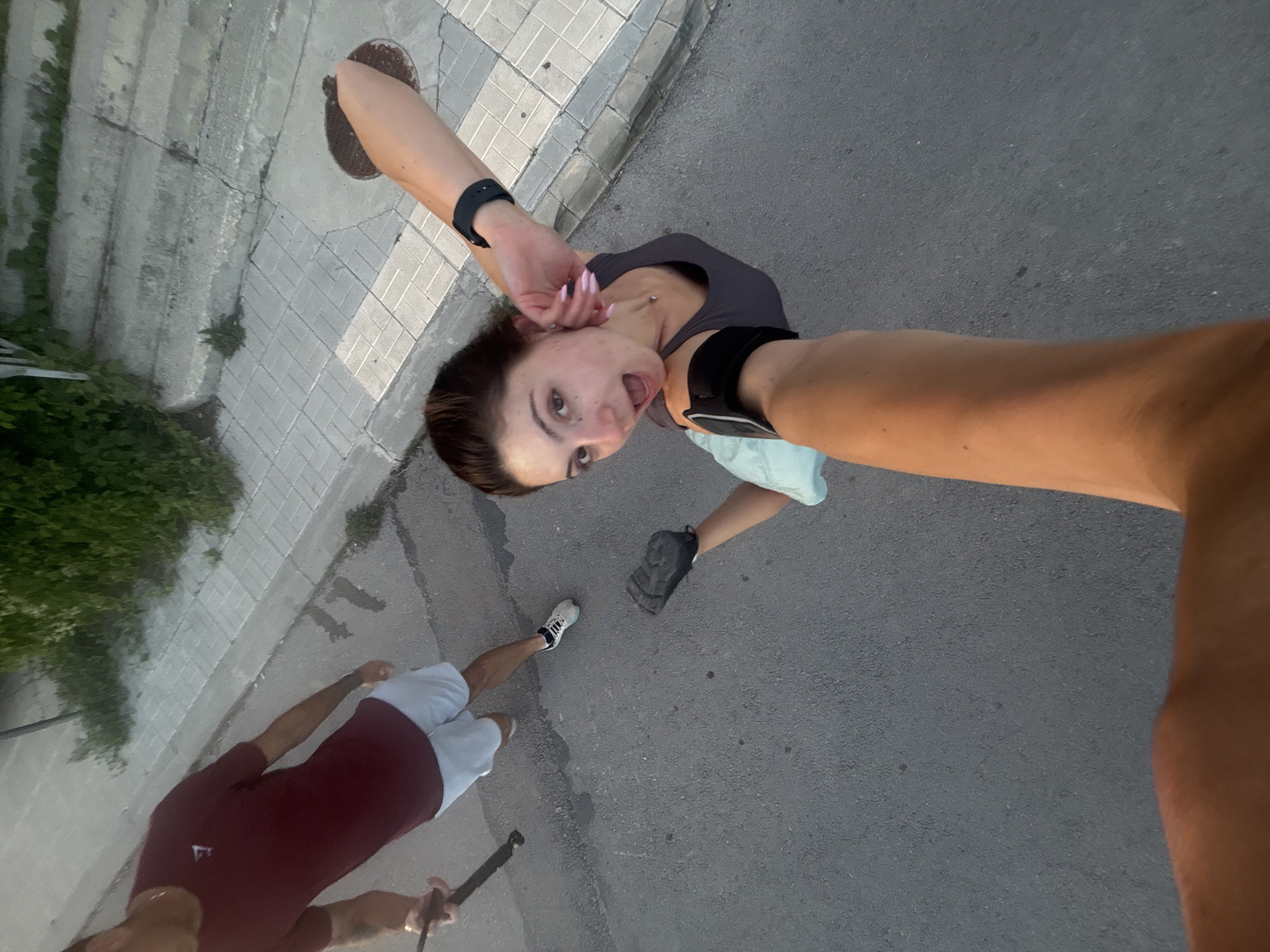 In Varna, Bulgaria, a couple is captured in a high-angle, selfie-style shot on a paved street next to a sidewalk with a manhole cover and concrete steps. The woman, in the foreground, is wearing a grey tank top and appears to be jogging or exercising. She is holding a phone or camera with her arm extended, looking surprised or excited with her mouth open. She has pink fingernails and a black fitness tracker on her left wrist. Her right arm is bent, and she is wearing a black fitness band on her upper arm. The man is in the background, wearing a maroon t-shirt and light-colored shorts. He is holding a black object, possibly a walking stick or phone mount, with his left hand. His right leg is bent as if he is walking or running. The environment suggests an outdoor urban setting with sparse greenery visible on the left side of the frame.