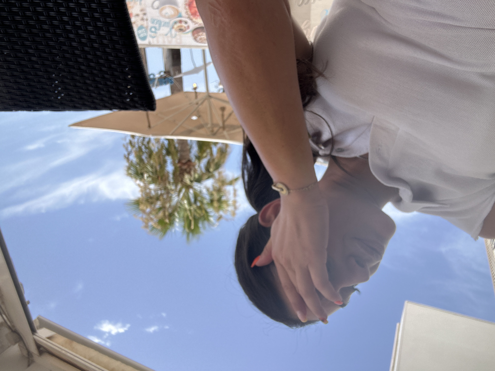 A woman is sitting outdoors at a cafe with a bright blue sky and scattered clouds. She is wearing a white polo shirt and her hand is covering her face. A palm tree and an open umbrella are visible in the background. A portion of a black woven chair is in the foreground. The scene is set in Cala Rajada, Spain.