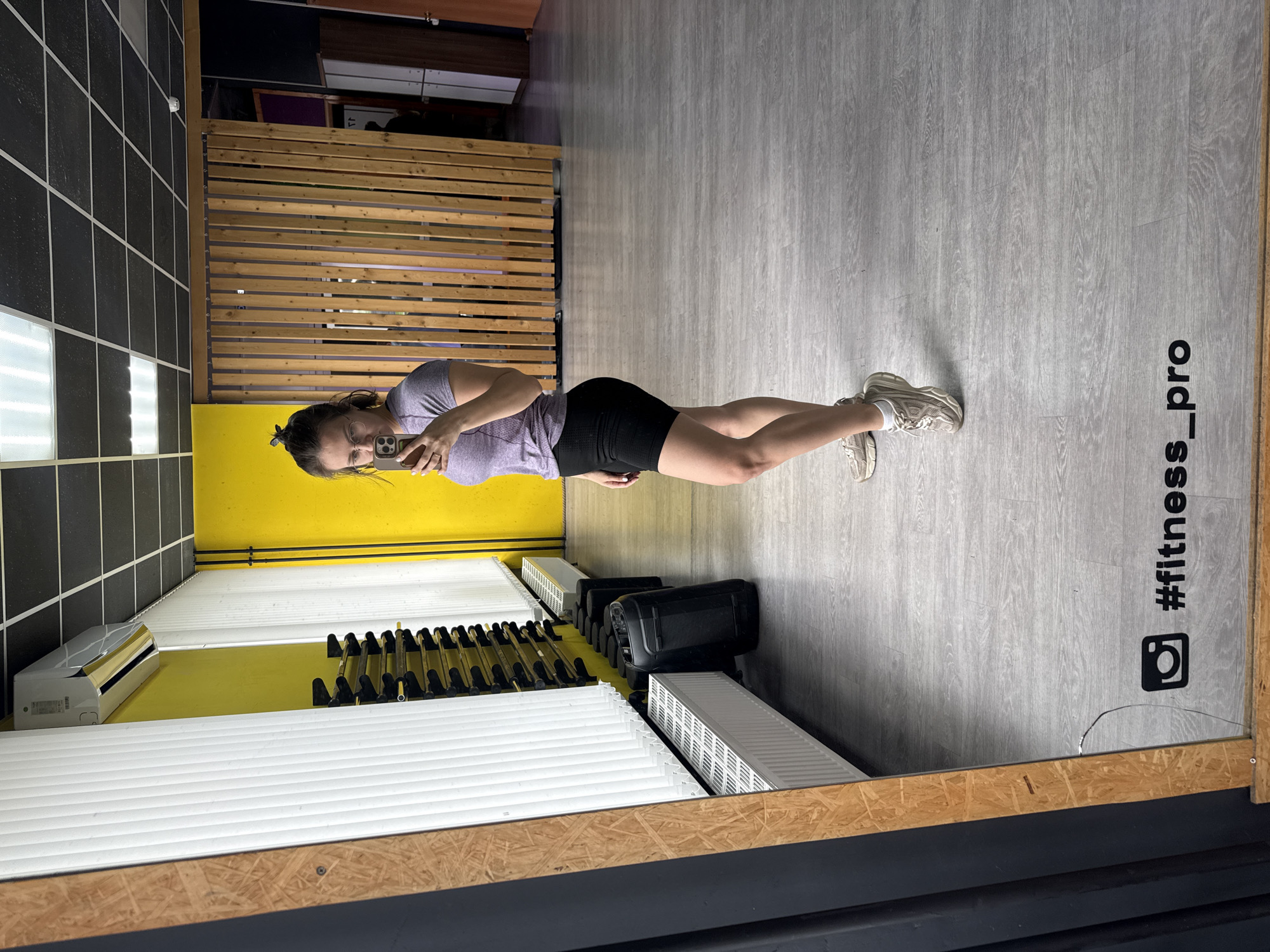 The image shows a woman taking a mirror selfie in a brightly lit fitness studio or gym. She stands upright, dressed in a light purple t-shirt, black athletic shorts, and light-colored sneakers with white socks, holding a smartphone up to her face.
The reflected environment reveals a gym setting. The floor is light grey with a wood-look pattern, and on it, an Instagram logo is visible along with the text "#fitness_pro". The walls feature a distinct design with bright yellow sections and areas covered in black tiles with white grout. A wooden slatted partition is also visible in the background.
Various gym equipment and utilities are reflected in the mirror: an air conditioning unit is mounted on a yellow wall, beneath which are several white, horizontal radiators or vents. Racks holding black handheld weights or kettlebells are lined up. A large black box-like object, possibly a speaker or other equipment, is also present. The space is illuminated by overhead fluorescent lights, some of which are visible in the reflection. The mirror itself has a wooden frame along its bottom and right edges, with a small crack visible in the bottom right corner. The overall scene suggests a functional and well-maintained fitness space.