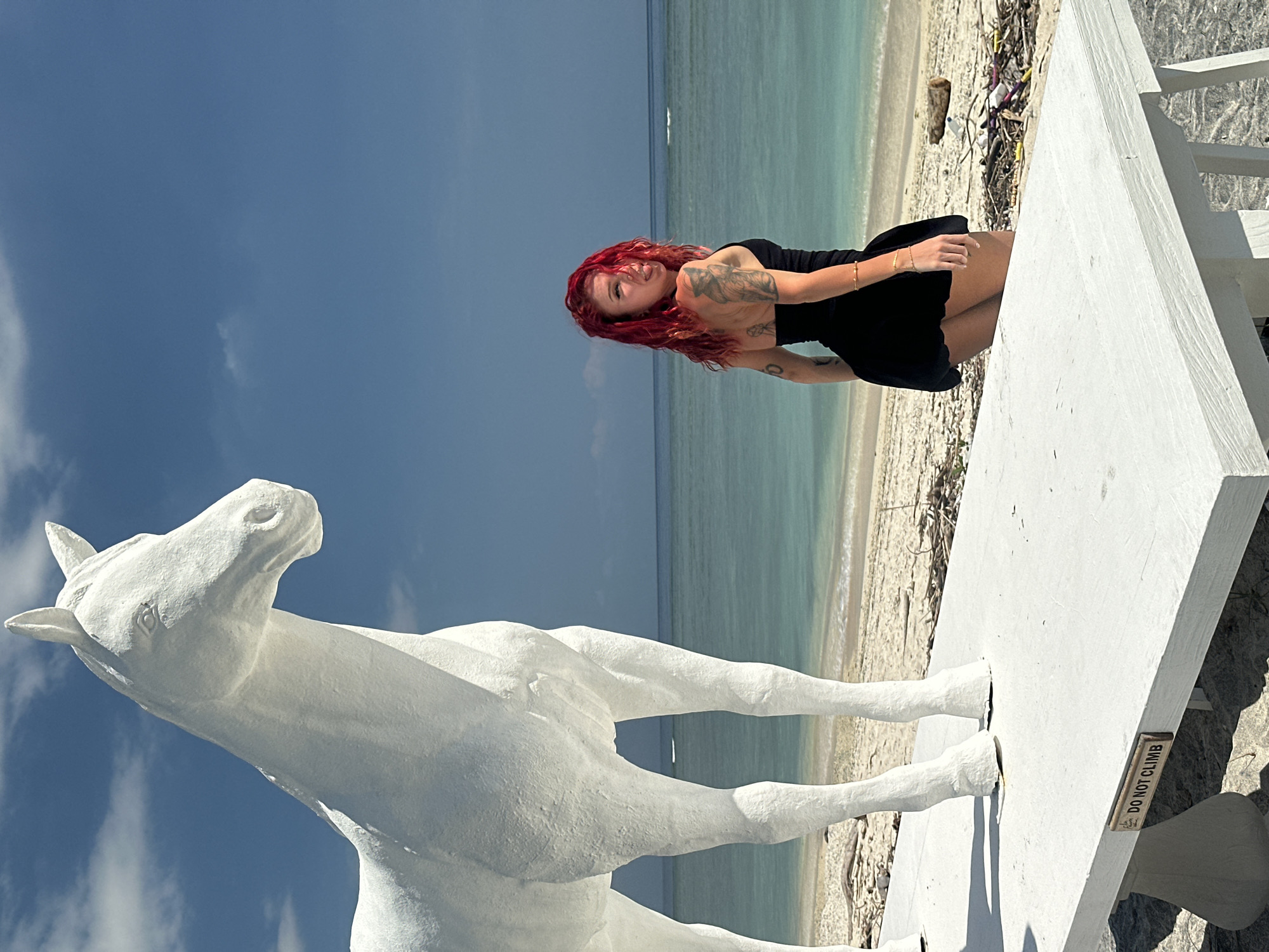 A full shot shows a red-haired woman near the coast in Unknown, Indonesia, next to a large white horse statue on a structure, with beach, ocean, and sky in the background. The sky has some scattered clouds. The woman is wearing a black dress and bracelets and has tattoos on her arms. She is positioned on the structure near the horse statue. The horse statue is white and appears to be made of concrete or plaster. The structure it stands on has a warning sign that reads, "DO NOT CLIMB." The beach and ocean stretch out behind the structure, and some debris is visible on the sand. The sky is blue.