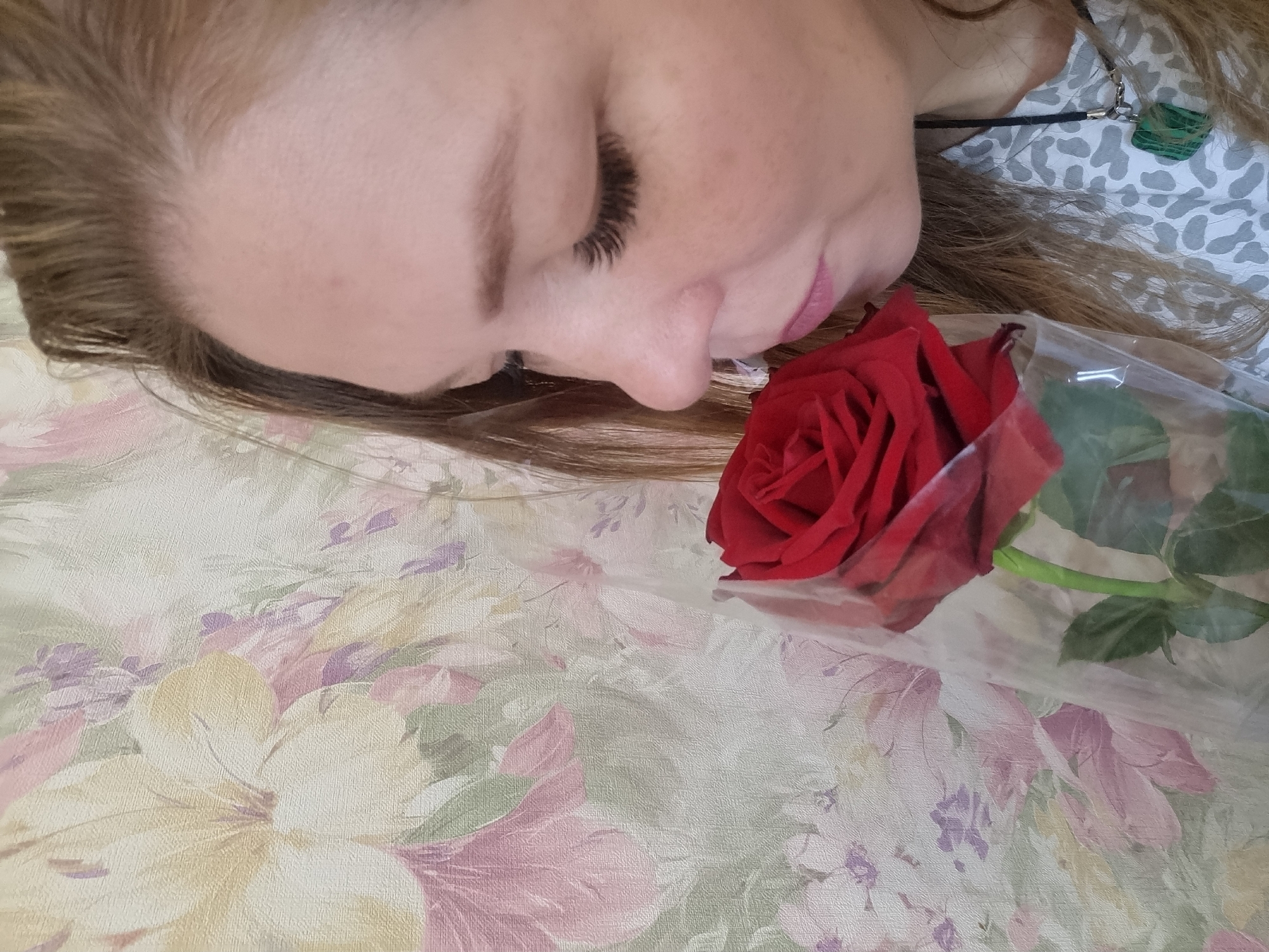 This close-up, high-angle image features a young woman lying down, her face captured from her forehead to just below her chin. Her eyes are closed, and she exhibits a serene expression, suggesting rest or contemplation. Her long, light brown hair frames her face, and she has pronounced dark eyelashes, likely false, along with light pink lipstick.
A vibrant red rose rests beside her face, near her mouth and chin. The rose is partially wrapped in clear plastic, with its green stem and some leaves visible through the film. Around her neck, a black cord necklace with a small, irregular green pendant (possibly a stone) is visible. She wears a light-colored top with a subtle, abstract grey and white pattern.
The woman and the rose are positioned on a soft, light-colored fabric, likely a bedspread or sofa cover, adorned with a muted floral pattern. This background features large blossoms and leaves in shades of cream, pale yellow, soft pink, purple, and light green. The overall mood conveyed is one of peacefulness and tender sentimentality. No text is visible, and the time of day or weather is not discernible. The scene is set indoors, in Chaikovsky, Russia.