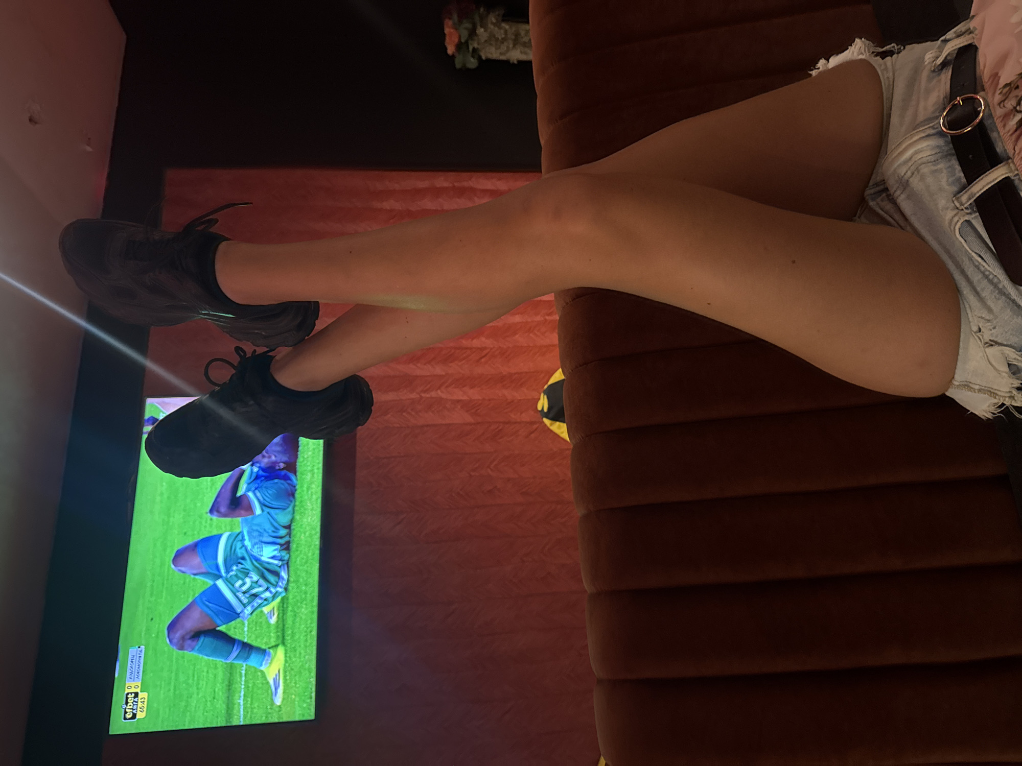 The image depicts a person sitting on a brown velvet couch, wearing denim shorts and black sneakers. The person's legs are crossed, with one foot near a television screen displaying a soccer match. The TV screen shows a soccer player in a green and blue uniform. The environment is dimly lit, and there is a flower arrangement in the background. The city and country of origin is Sofia, Bulgaria.