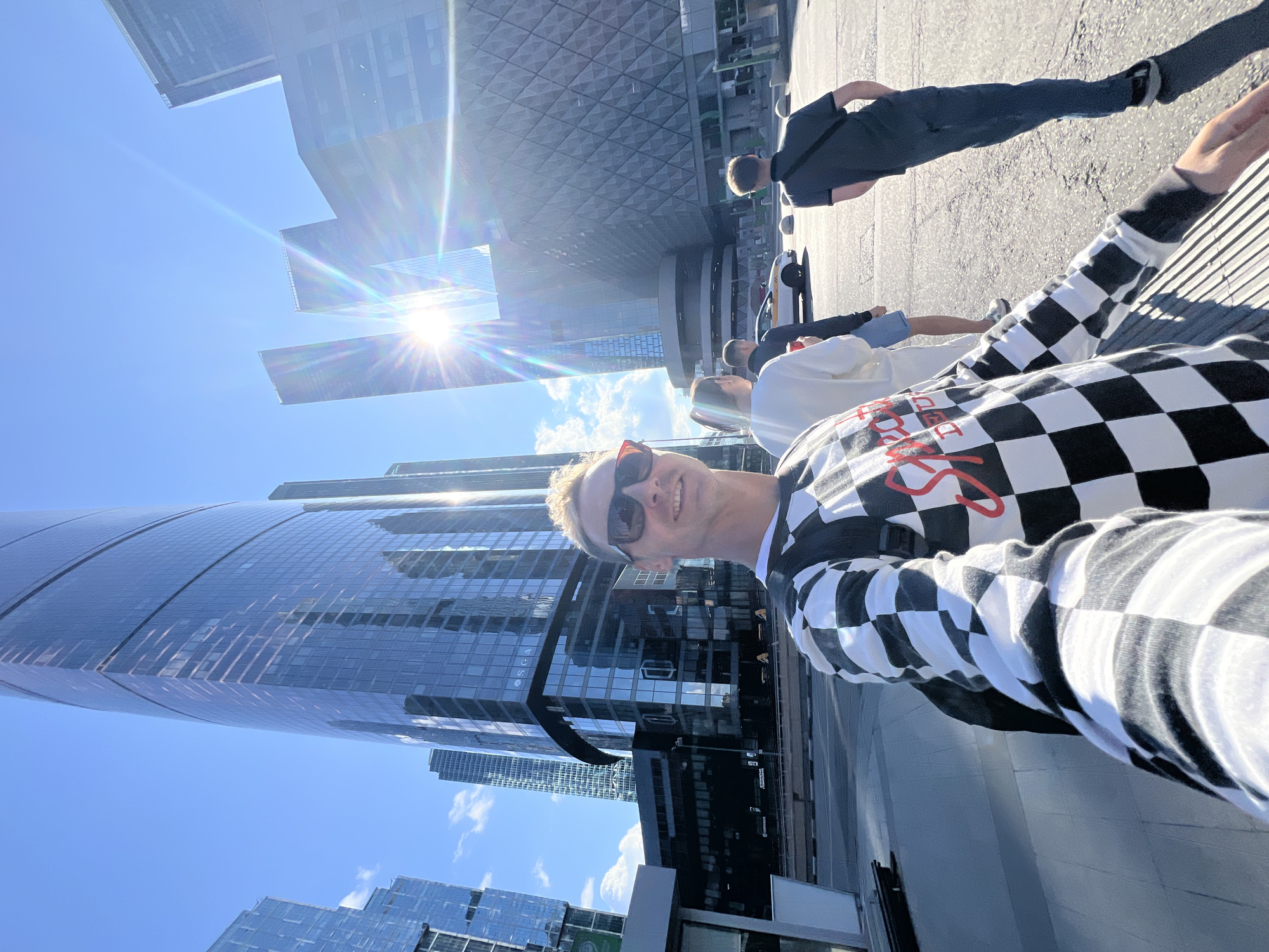 A man in a checkered long-sleeve shirt and sunglasses smiles at the camera while taking a selfie. Behind him, in the background, are modern skyscrapers and a bright sun shining through the sky. Several other people are walking in the background. The location is the Moscow City business district in Khimki, Russia.