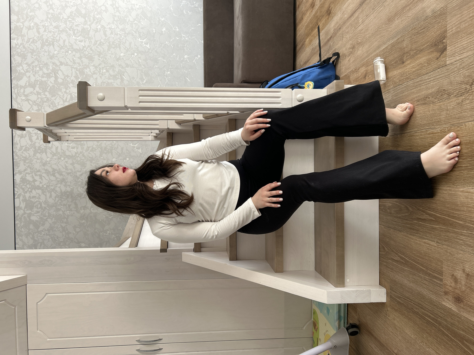 The image depicts a young woman sitting on wooden stairs inside a home in Chaikovsky, Russia. She has long, dark brown hair and is wearing a long-sleeved white top and black flared pants. Her fingernails and toenails are painted with light-colored polish, and she has red lipstick. She is seated on the fourth step from the bottom, looking upwards and slightly to her left with a calm or pensive expression. Her arms are resting on her knees in a relaxed posture.
The setting is a modern indoor residential space. The stairs are white wood with decorative balusters and a railing. The floor throughout the visible area is a light brown wood-look material. In the background, there is a wall with light grey marbled wallpaper and a brown sofa. On the floor near the stairs, a blue backpack with yellow accents and a clear plastic jar with a white lid are visible. To the left, beneath the stairs, a white cabinet with drawers and pull handles is present. A colorful mat and part of a white stand or appliance can also be seen on the floor. The ambient lighting suggests it is daytime. No strong emotions are conveyed, and no visible text is present.