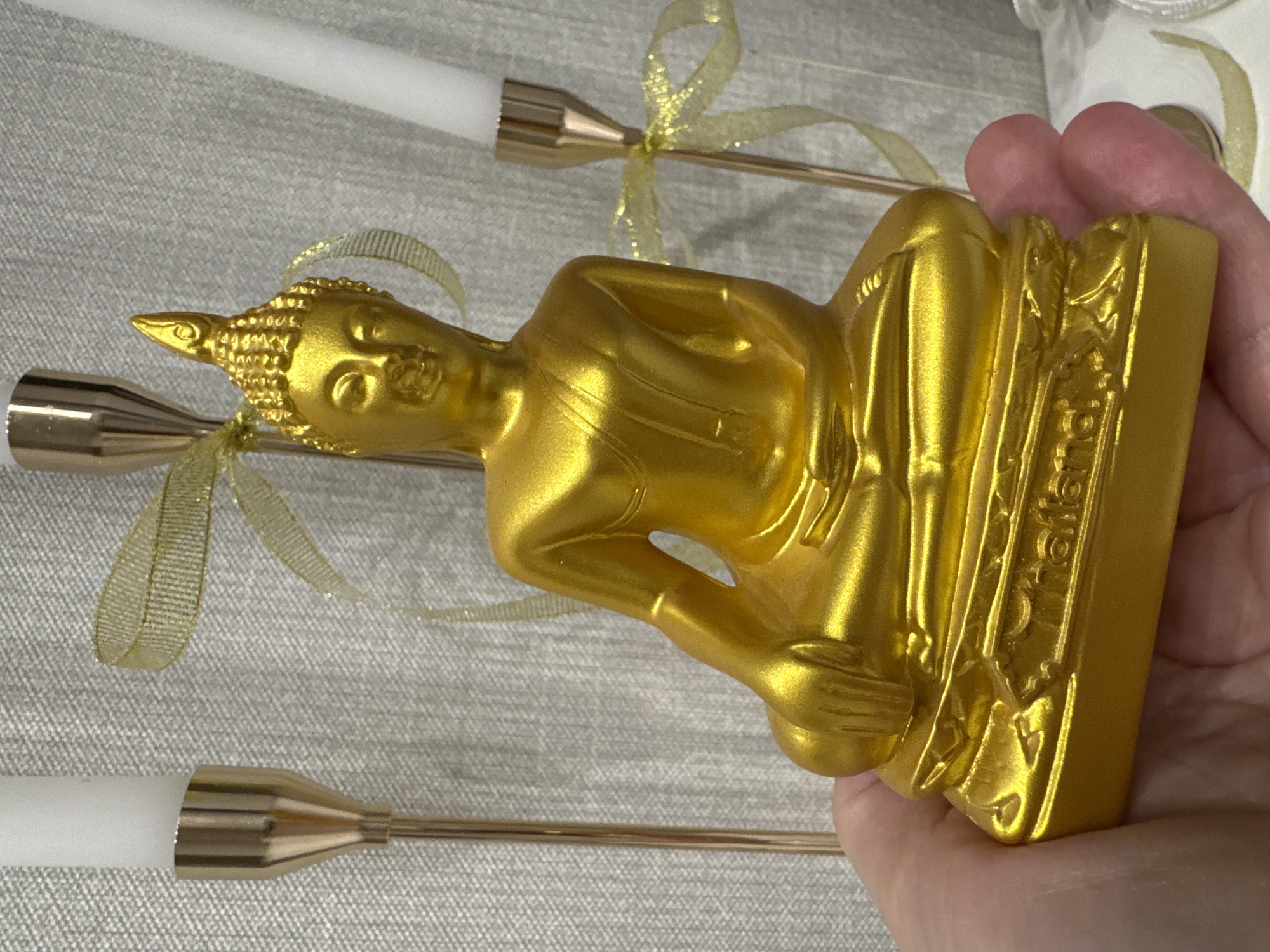 A person's hand gently holds a small, golden Buddha statue in a meditative pose. The statue is richly golden and appears to be made of metal or a metallic-coated material. On the base of the statue, the word "Thailand" is distinctly visible, along with some less legible decorative script above it.
The statue is positioned vertically in the image, suggesting it is being held upright. In the background, partially out of focus, are two white taper candles set in elegant, slender golden candle holders. Each candle holder is adorned with a delicate, shimmering golden ribbon tied into a bow, adding a decorative touch. The candles and their holders are arranged against a light gray, subtly textured fabric surface, possibly linen or a similar material, which serves as the backdrop. The lighting is bright and even, highlighting the golden sheen of the statue and candle holders. The scene appears to be an indoor setting, likely a display or arrangement, possibly for an event or as a decorative item in Chaikovsky, Russia.
