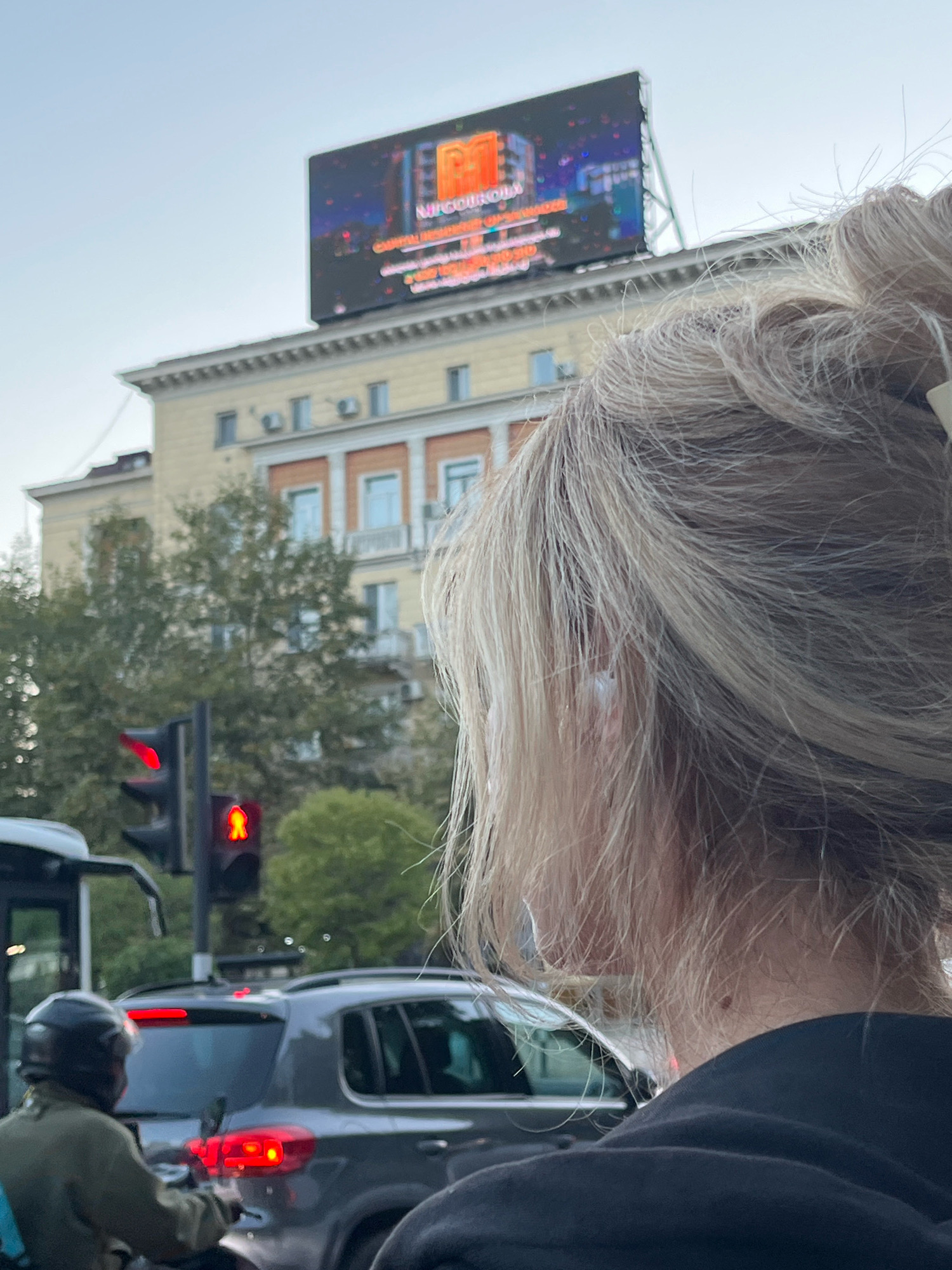 The image shows a close-up view of a woman's head and shoulders from behind, with her blonde hair pulled up. In the background, a multi-story building with light yellow facade and orange accents is visible. Above the building, a large digital billboard displays an advertisement with colorful graphics, including what appears to be a building and the text "NITROKIDS" in large red letters. A traffic light with a red signal and a red pedestrian crossing signal are present. Several vehicles, including a gray SUV and a scooter with a rider wearing a helmet, are on the road. The setting appears to be a city street in Tbilisi, Georgia, as indicated by the architecture and the style of the billboard.