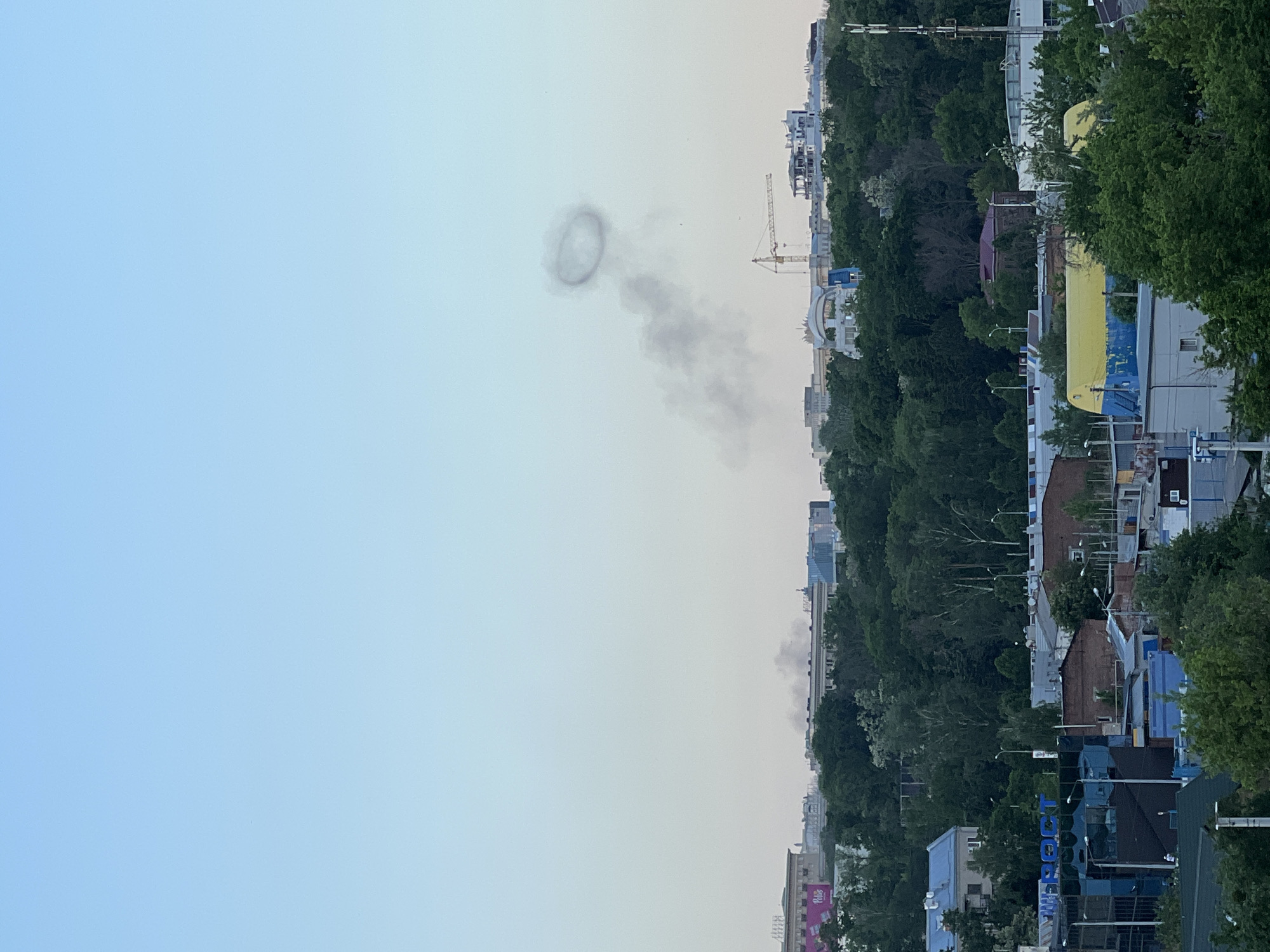 The image shows a cityscape under a clear sky. A distinct ring of smoke is visible in the sky. The city has a mix of high-rise buildings and lower residential structures, with significant tree cover. A construction crane is present in the background. The scene suggests an urban environment, but the precise location within the city is unclear.
