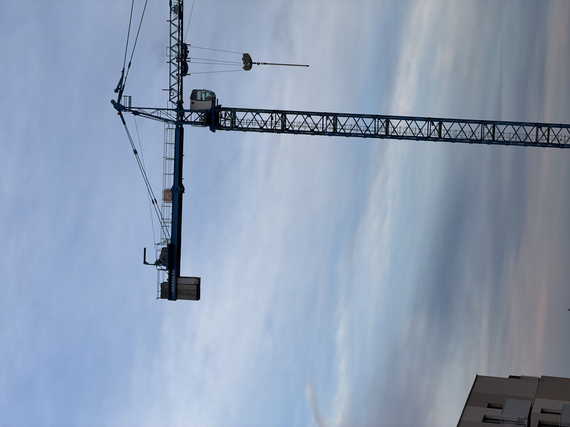A tall blue construction crane is silhouetted against a cloudy sky in Łódź, Poland. The crane's horizontal jib extends to the left, with counterweights visible at the far end and a cabin for the operator situated on the vertical mast. A hook hangs from the jib. In the lower left corner of the image, the corner of a modern, multi-story building with white balconies is visible. The overall scene suggests an ongoing construction project in an urban environment.
