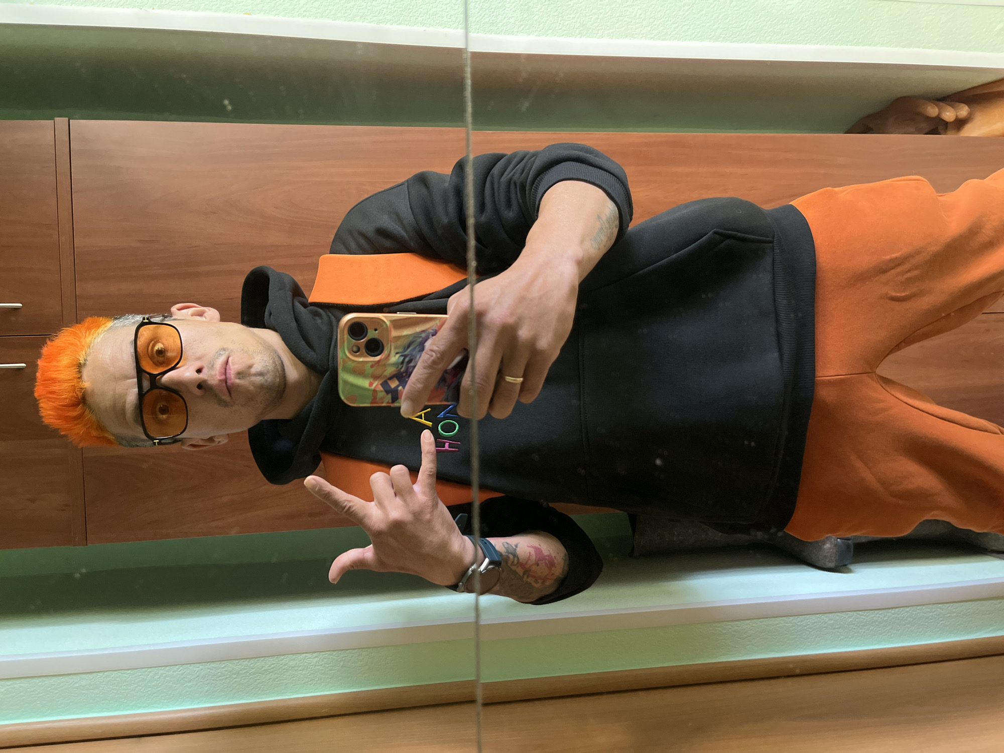 A man takes a selfie in a mirror. He is wearing orange hair, orange-tinted glasses, a black hoodie with an orange shoulder strap, and orange sweatpants. He is making the "rock on" hand gesture. The reflection in the mirror shows a light green wall, a wooden headboard, and a glimpse of a sculpture of hands on a shelf. The phone case has colorful letters spelling "HELLO". The photograph may have been taken in the city of Chaikovsky, Russia.