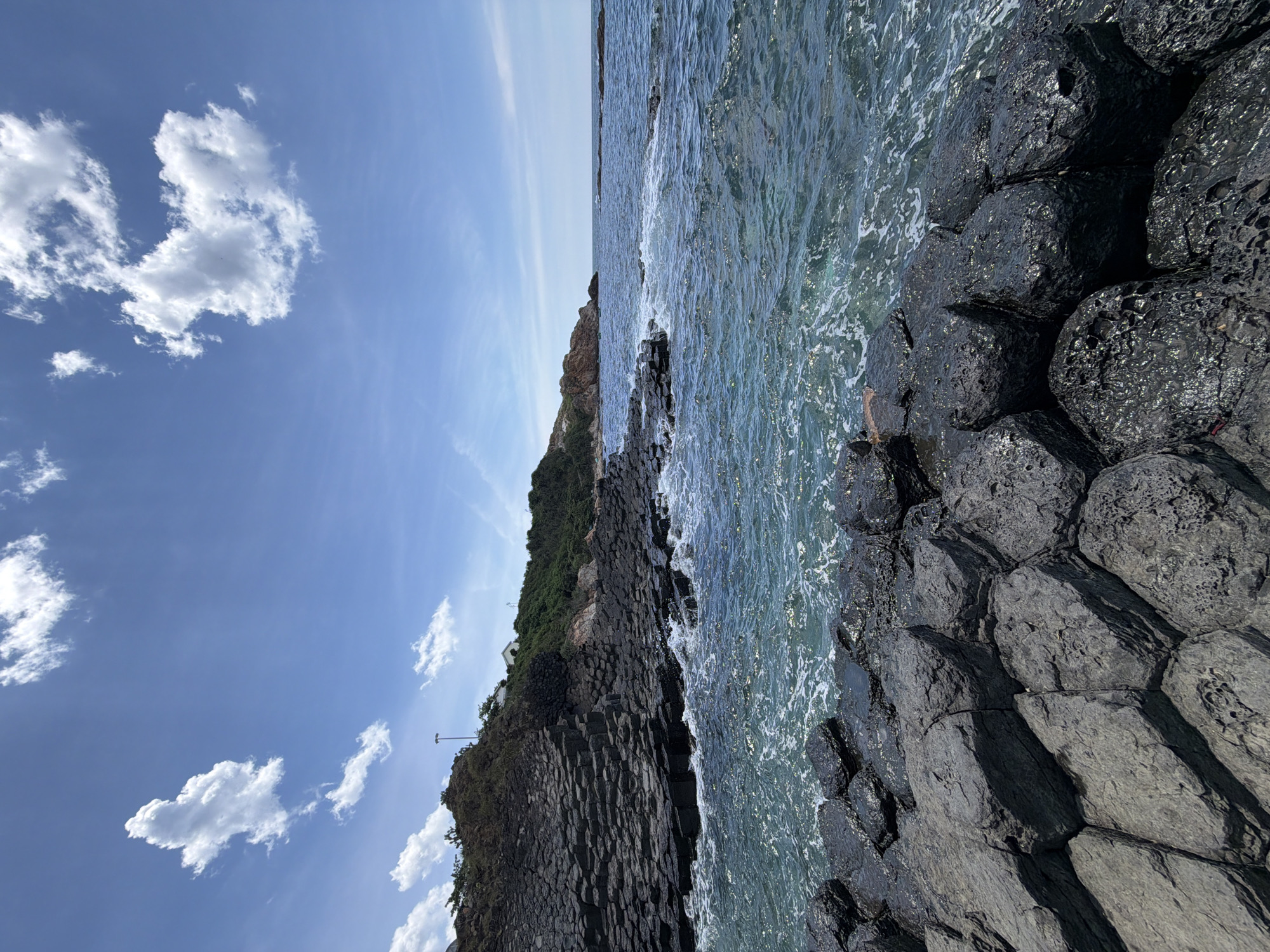 The image depicts a coastal scene at Phú Lương, Vietnam. The foreground is dominated by dark, hexagonal basalt rock formations that jut out into the turquoise ocean. These rocks are weathered and show a textured surface. The water is choppy with white foam where it meets the rocks. In the mid-ground, more of these hexagonal rock formations continue along the coastline, leading up to a green, vegetated hillside. White buildings and a lamppost are visible on the crest of the hill. The sky is a bright blue with scattered white clouds. The overall impression is one of a rugged, natural coastal landscape.