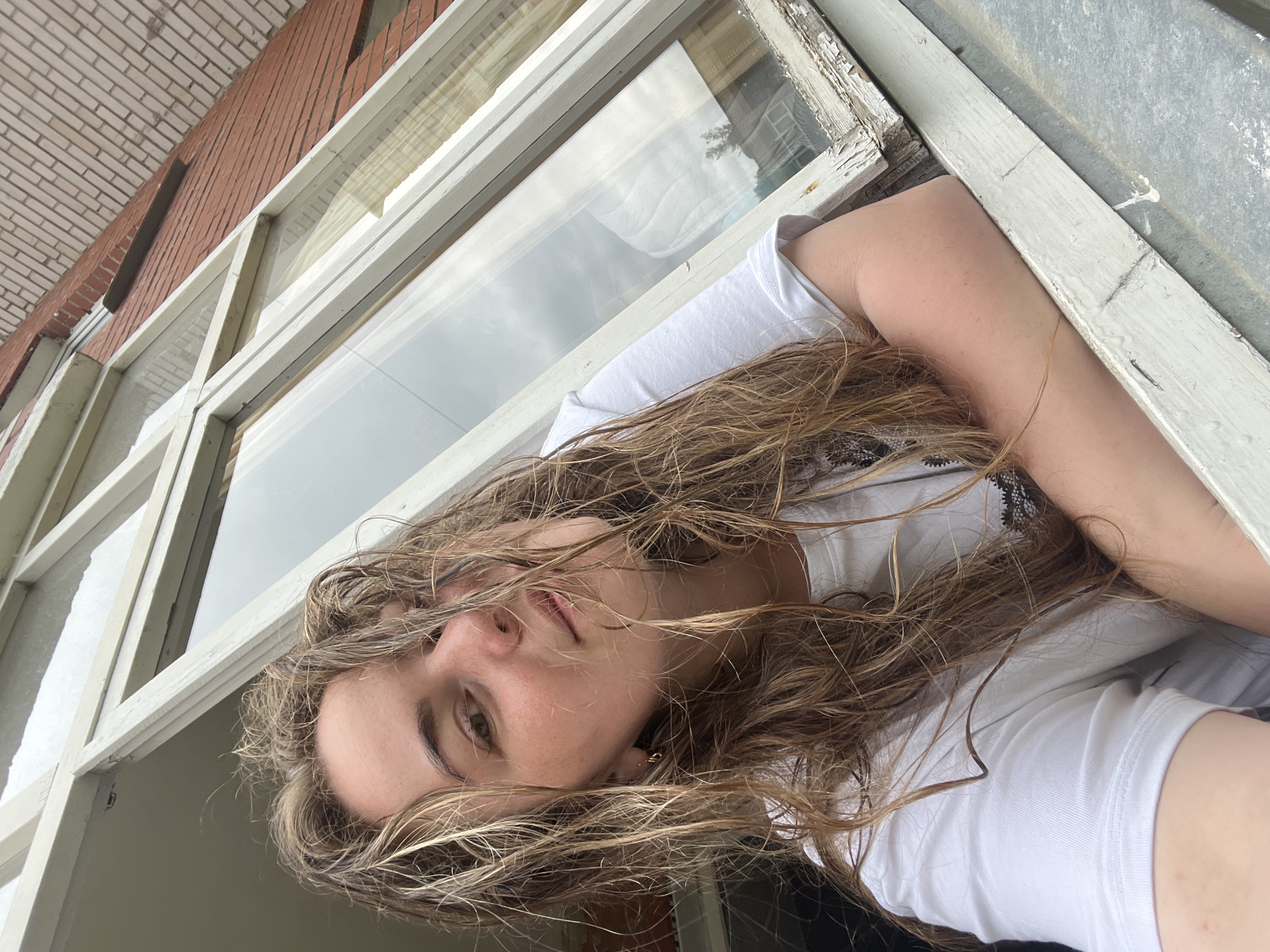 A young woman with long, wet, blonde hair is leaning out of an open window. She is wearing a white t-shirt. The window frame is white and appears to be old and weathered. Behind her, a brick building is visible. The sky is overcast. The image appears to be taken from a low angle, looking up at the woman. This scene is not located in the city of Chaikovsky, Russia.