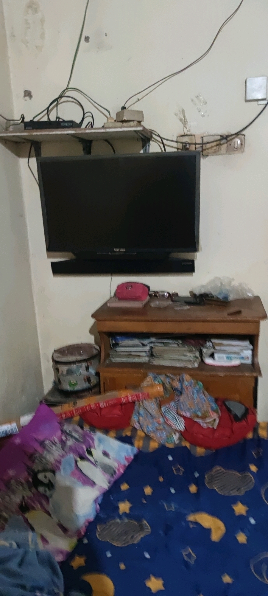The image depicts a close-up, ground-level view of a cluttered bedroom in Gambarsari, Indonesia. A black flat-screen television is mounted on the wall above a wooden shelf unit. The shelf unit holds various items, including books, a pink cosmetic bag, and a cylindrical object that appears to be a rice cooker. The floor in front of the shelf is covered with a blue bedsheet adorned with yellow stars and clouds, and piles of colorful clothing and bedding. Electrical cords and power outlets are visible on the wall behind the television. The overall scene suggests a lived-in, perhaps slightly disorganized, domestic space.