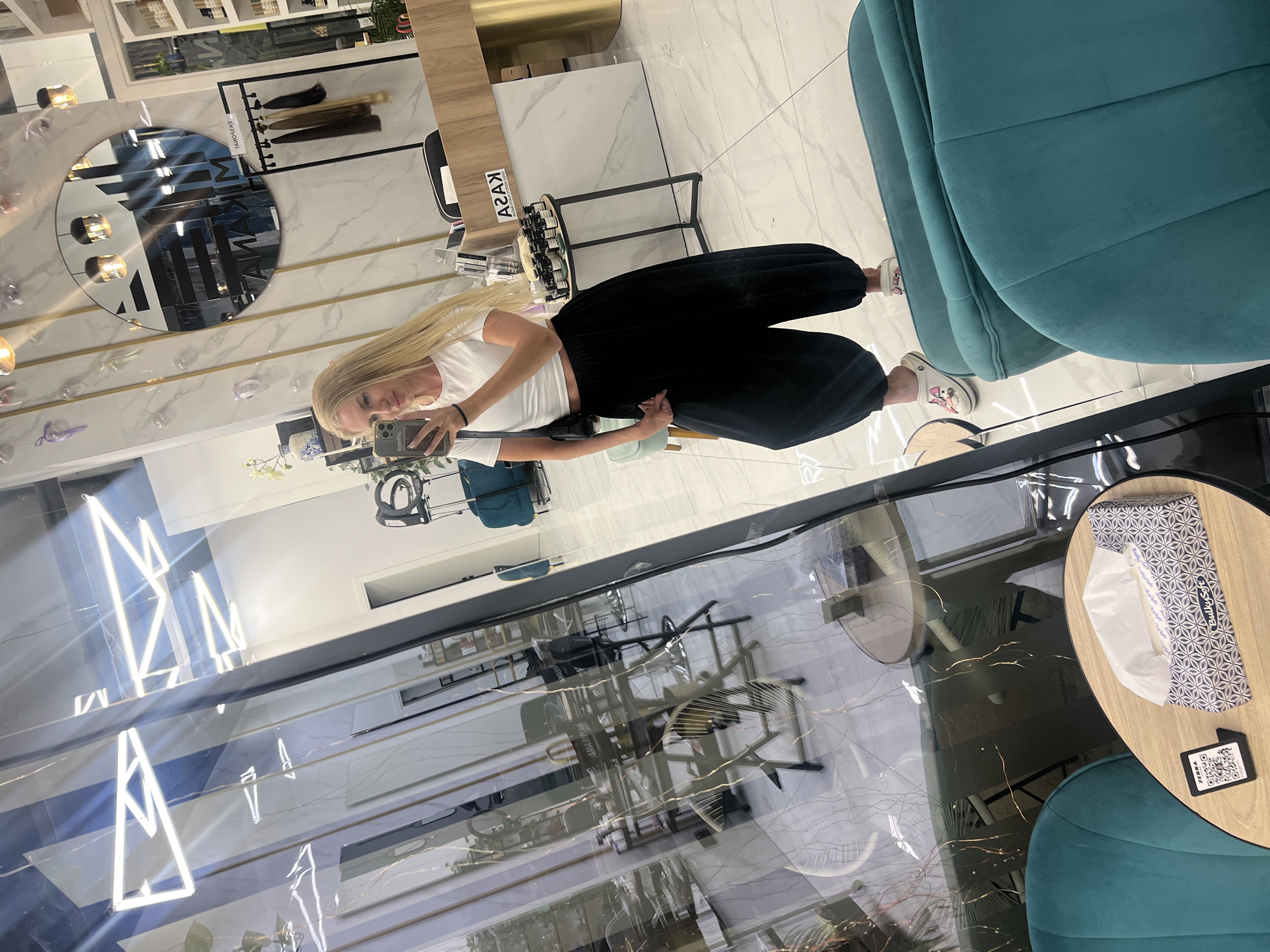 A woman with long blonde hair is taking a selfie in a salon in Budva, Montenegro. She is wearing a white crop top and black voluminous pants, with white clogs on her feet. She has a black purse slung over her shoulder. The salon has a modern design with marble walls, gold accents, and geometric lighting fixtures. In the background, there is a round mirror reflecting the salon interior, a rack of hair extensions, shelves stocked with products, and a turquoise sofa. The floor is tiled white.