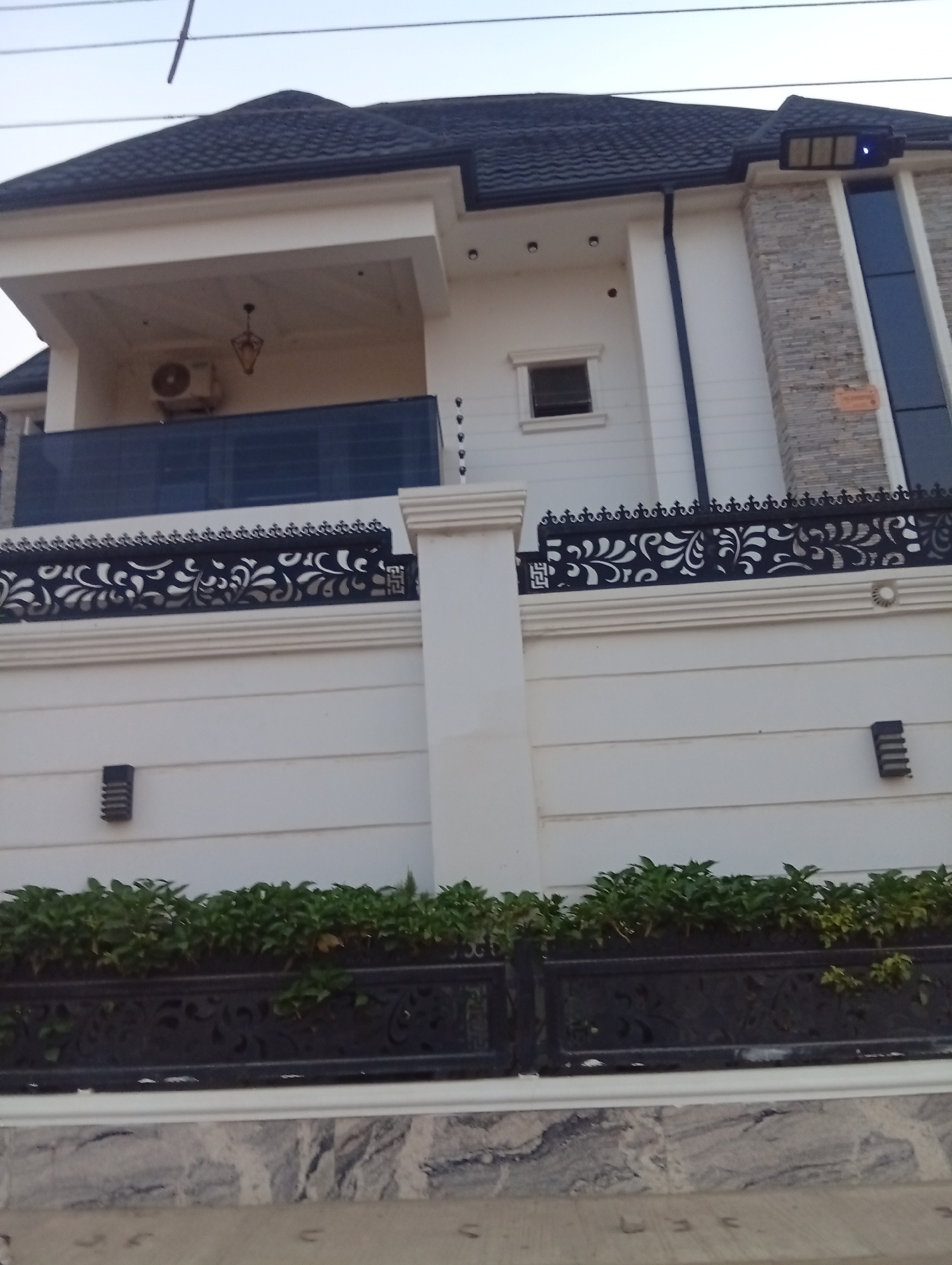The image depicts a modern-style, two-story building located in Aviation Quarters, Nigeria. The building features a dark, tiled roof, white exterior walls, and stone detailing on a vertical section to the right. A balcony is visible on the second floor, with a dark glass railing and an air conditioning unit installed on the wall behind it. The property is enclosed by a white wall with decorative black metalwork along the top. Green plants are situated in front of the wall. The ground in the foreground appears to be paved with gray and white patterned tiles. Several electrical wires are visible against the sky, suggesting an urban or suburban setting.