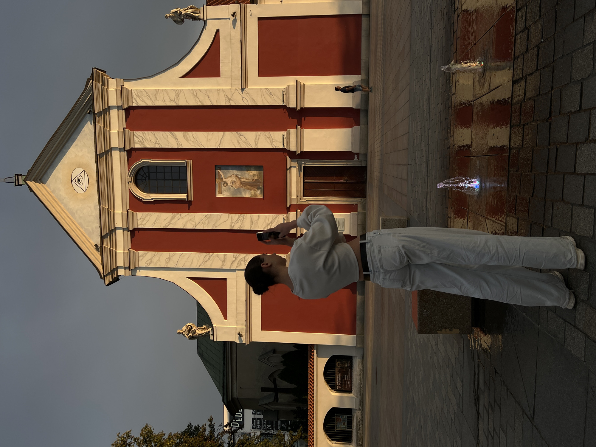 A young woman, viewed from behind and facing away from the camera, stands in a paved square in front of a red and white building with architectural details. She is wearing a white cropped sweatshirt and loose-fitting light blue jeans, and is holding a smartphone to take a picture. The building appears to be a church, with a prominent pediment featuring an eye within a triangle symbol and a painting of a religious scene above a large wooden door. Two statues are visible on the facade, one on each side of the pediment. A lone man in a dark t-shirt and shorts is walking in the distance. In the foreground, two small, illuminated fountains spray water onto the wet pavement. The scene is illuminated by warm, low-angled sunlight, suggesting it is either early morning or late afternoon. The paving stones of the square are visible in detail, with some appearing wet. There are also some concrete structures in the foreground. In the background, a portion of another building with a green roof and arched windows can be seen, with signage that includes the word "DE LUXE". This location is the Holy Spirit Church in Lublin, Poland.