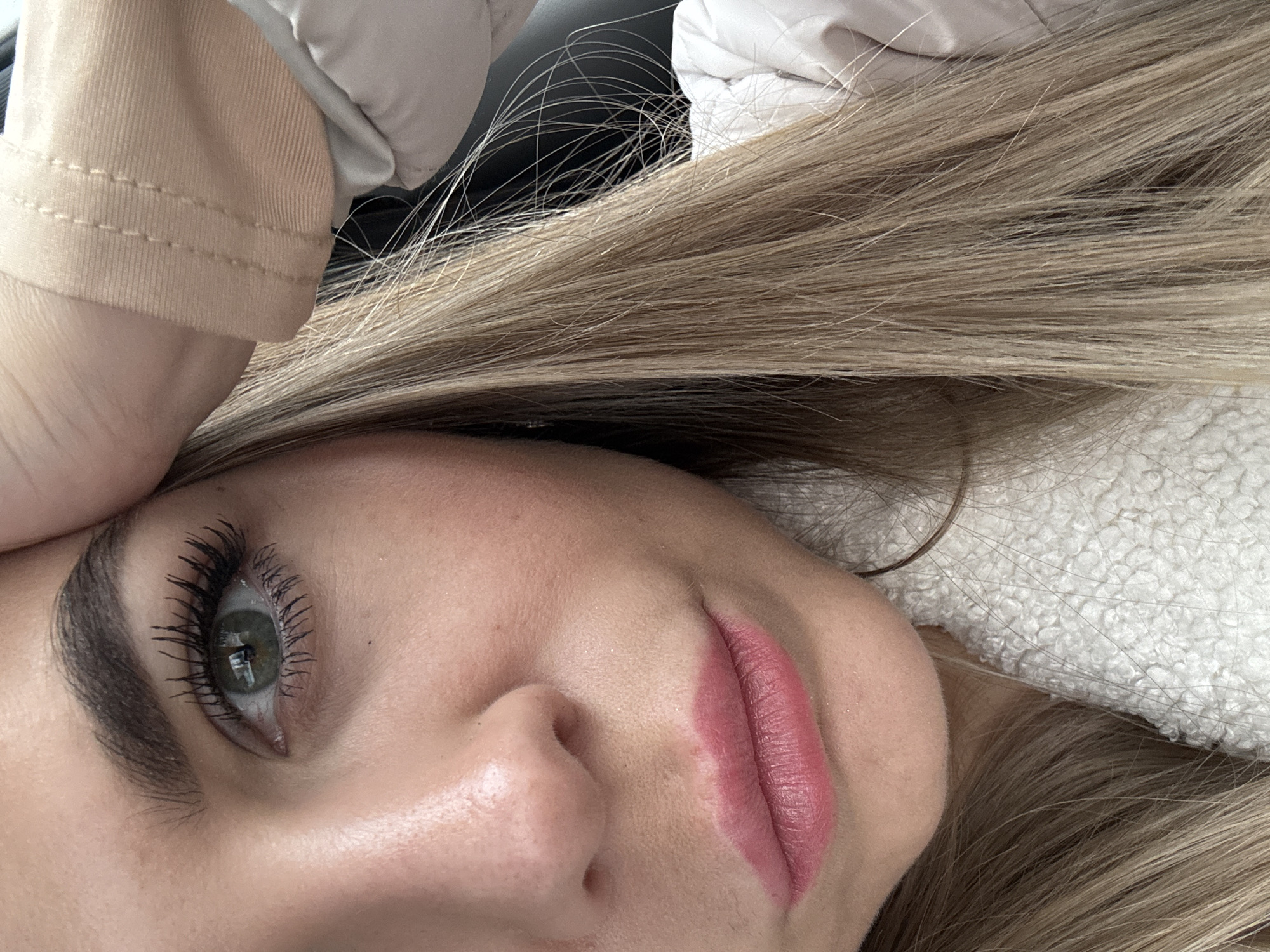The image depicts a close-up, angled portrait of a female subject in the city of Chaikovsky, Russia. Approximately half of her face is visible, from above the left eyebrow to below the chin, with her head slightly tilted. Her left eye is prominent, featuring a light green or hazel iris with a dark pupil and long, dark, mascara-enhanced eyelashes. The eyebrow is dark brown and well-defined. Her skin exhibits a fair complexion with a subtle rosiness on the cheek and minimal texture visible. A small, dark mark is present below the left eye. Her lips are full and coated with a reddish-pink lipstick. Long, straight to slightly wavy blonde hair with lighter streaks extends downwards on the right side of the frame. She wears a light beige or tan garment with visible stitching near her right temple, possibly a sleeve or collar, and a white, textured 'sherpa'-style material garment is visible at the bottom right. The background is dark and out of focus, providing no specific environmental details. The subject's right hand or forearm is positioned near her right temple.