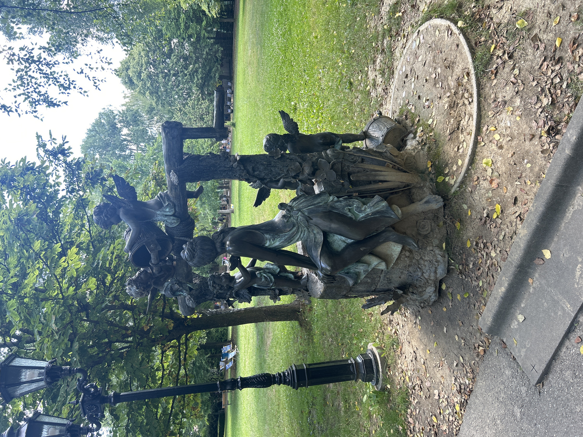 The image depicts a bronze sculpture of cherubic figures and a woman in a park setting. A dark, ornate lamppost stands to the left of the sculpture, which is surrounded by a grassy area with scattered leaves. In the background, more trees are visible, along with lounge chairs and a fence. The scene is likely outdoors in a public park, given the presence of the sculpture, lamppost, and manicured lawn.