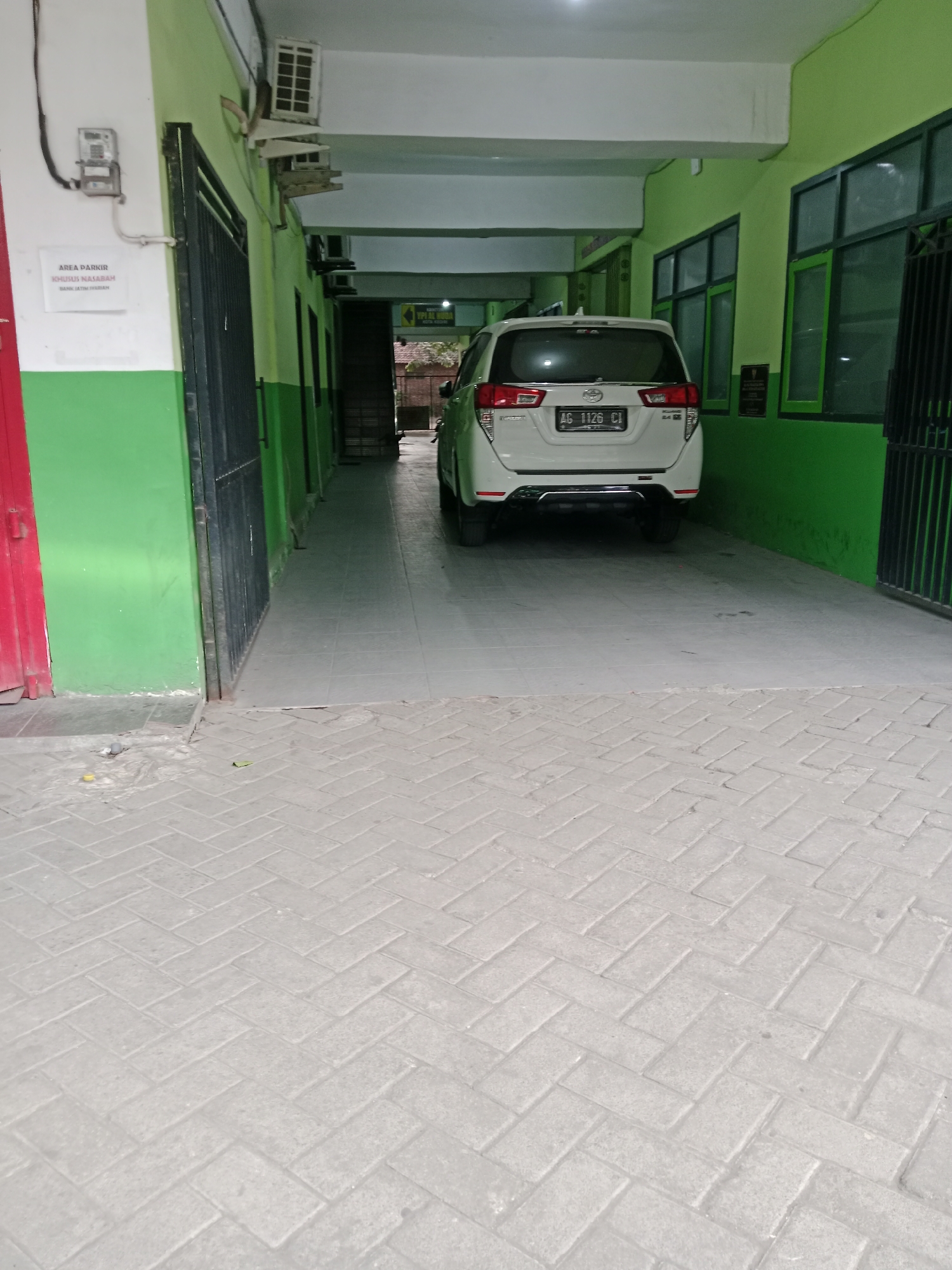 The image captures a white SUV parked in an outdoor covered area, likely a parking lot or a driveway. The vehicle has a license plate that reads "AG 1126 CI". The surrounding environment features green painted walls and a grey tiled floor. A sign is visible on the wall to the left of the vehicle, with text that appears to be in Indonesian, indicating "AREA PARKIR KHUSUS NASABAH BANK JATIM SYARIAH" (Parking area for Bank Jatim Syariah customers). The overall scene suggests a commercial or financial establishment in Kediri City, Indonesia, specifically a bank with dedicated parking for its customers.