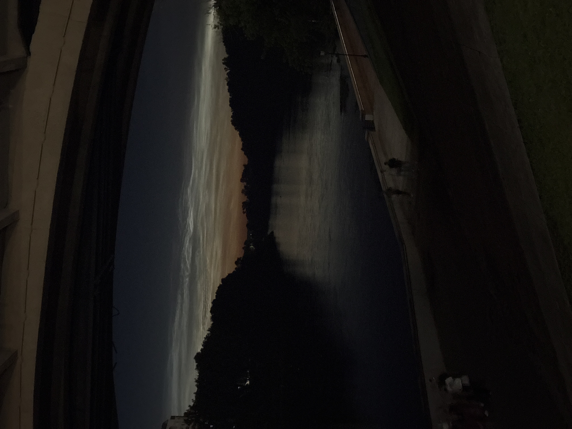 The image depicts a wide river at dusk, with trees silhouetted along the banks. The sky above the trees is dark with faint, luminous clouds. The foreground shows a paved walkway bordering the river, with a few dimly lit figures walking along it. A grassy slope is visible in the bottom right corner. The scene is observed from beneath an overpass or bridge. The location is the Western Dvina River in Vitsebsk, Belarus, viewed from under the bridge on Lenin Street.