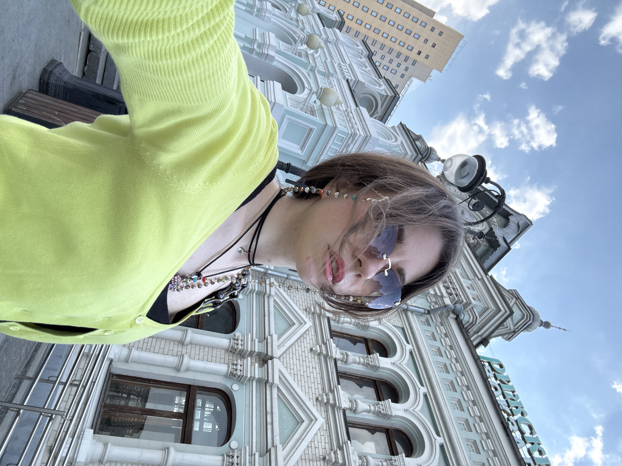 A young woman wearing a yellow cardigan and sunglasses is taking a selfie in front of the TSUM department store in Moscow, Russia. The building's ornate facade, with its arched windows and decorative elements, is prominently visible in the background. Above the TSUM building, a modern building and a blue sky with scattered clouds can be seen. The woman is wearing multiple necklaces and what appears to be a beaded eyeglass chain.