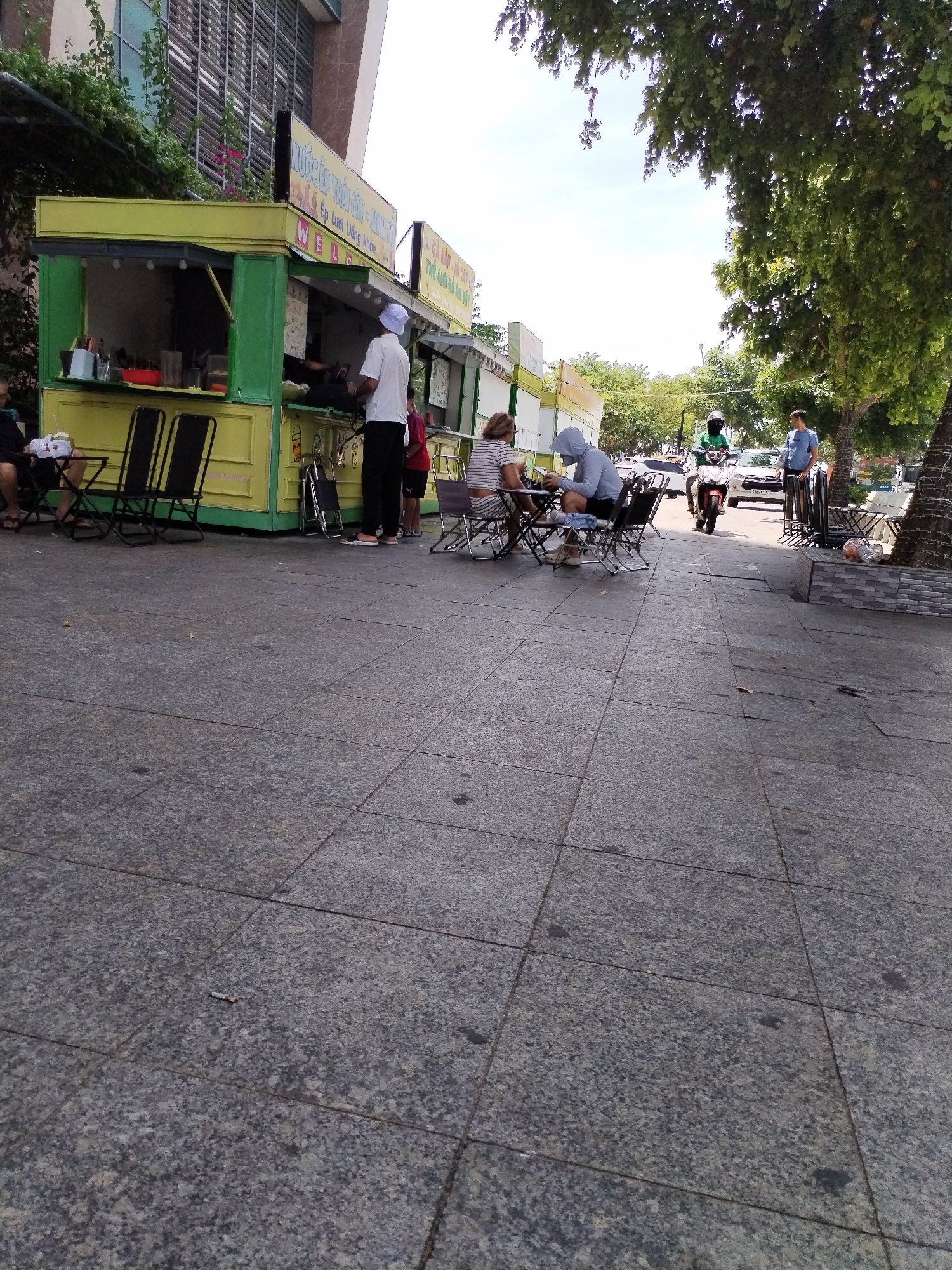 The image depicts a street-level view of a food stall in Nha Trang Ward, Vietnam. The stall, painted bright yellow and green, has an open front displaying food preparation areas. Several black metal folding chairs are set up outside the stall, with some occupied by patrons. One person, wearing a white shirt and hat, stands behind the counter, seemingly serving customers. Two individuals, one in a striped shirt and another in a hooded gray sweatshirt, are seated at a table. In the background, a scooter rider in a green shirt and helmet navigates the street, with a white car and other vehicles visible further down. Lush green trees canopy the scene, suggesting an outdoor urban setting. The ground is paved with large, grey granite tiles.