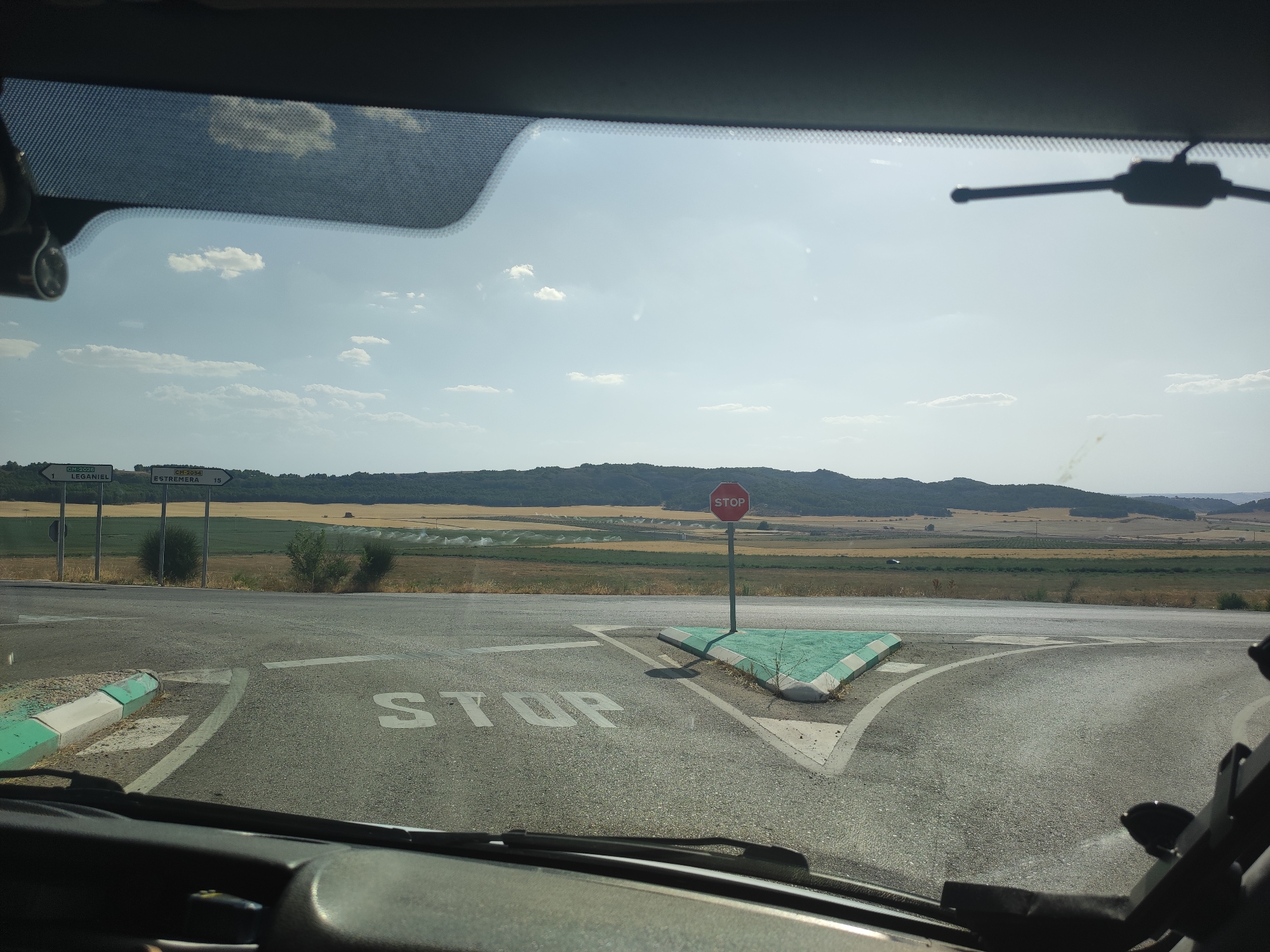 The image is taken from inside a vehicle, looking out through the windshield at a rural intersection. The road surface is asphalt with white painted lines indicating lanes and a stop line. A stop sign, octagonal and red with white lettering, is prominently displayed on the right side of the intersection. The ground around the stop sign is a raised triangular island with green and white striped edging. On the left side of the intersection, there are two white directional signs. One points left and indicates "CM-2020 LEGANIAL 1", and the other points right and indicates "CM-2054 ESTREMERA 15". Beyond the intersection, the landscape consists of rolling hills with cultivated fields in various shades of green and yellow, and a distant treeline. The sky is bright blue with scattered white clouds. The image appears to be taken in the vicinity of Leganiel, Spain, as indicated by the road signs.