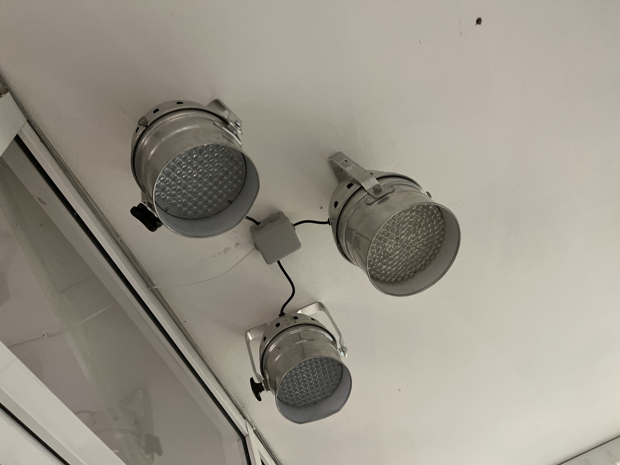 The image shows three metallic, cylindrical LED spotlights mounted on a white ceiling. Each spotlight has a textured, reflective front panel composed of numerous small, hexagonal lenses. Two of the spotlights are positioned on the left side of the image, with one slightly above and to the left of the other. The third spotlight is located to the right and slightly above the two on the left. A small, gray electrical junction box is visible between the top-left and the right spotlight, with black wires connecting to the lights. Above the spotlights, a reflective, white surface, possibly a window or a skylight, is visible, showing some indistinct reflections. The overall environment appears to be indoors, likely a room or a studio. There are no people or any clear indications of the city of Ivanovo, Russia.