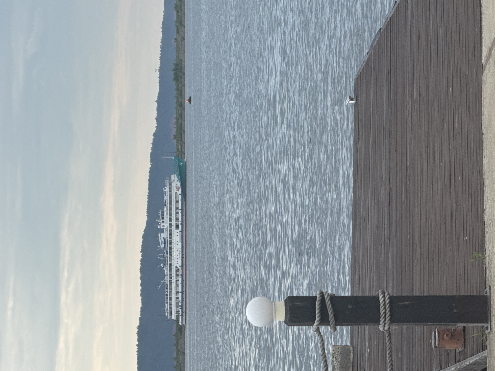 An exterior, eye-level, medium shot shows a large white riverboat with multiple decks and a turquoise hull in the distance, sailing on a wide body of water. The boat has numerous windows and railings, and its name appears to be "Volga-Don." In the foreground, a wooden pier extends into the water. A dark wooden post with a white spherical light fixture and thick ropes wrapped around it stands on the pier. The water is rippled, and a small red and orange boat with a person in it is visible in the mid-ground. The background features a dark, forested hillside under a cloudy sky. The scene is set on the Kama River in Chaikovsky, Russia, a city known for its river port and hydroelectric power station.