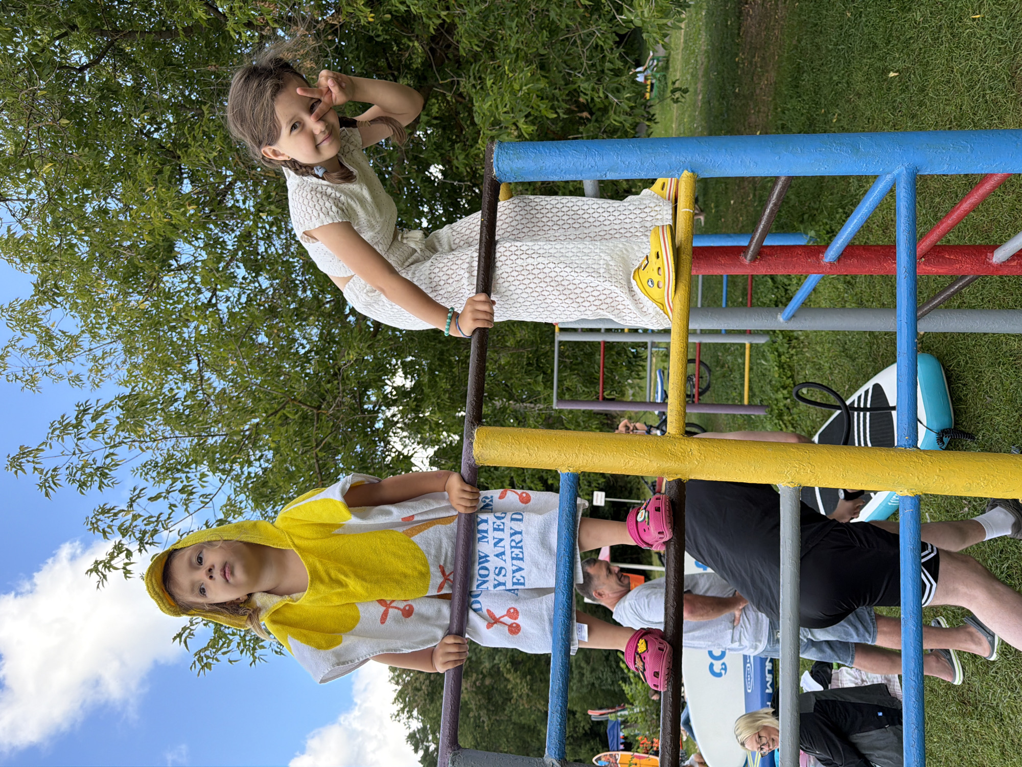In Соколово, Russia, two young girls are on a playground structure. The girl on the left, wearing a yellow hooded towel with a cherry pattern and pink Crocs, is looking up at the sky. The girl on the right, dressed in a white lace outfit and yellow Crocs, is leaning forward and making a peace sign with her fingers, looking at the camera. They are on metal bars of a playground. In the background, there are trees, a blue sky with clouds, and other people and objects, including what appear to be paddleboards and a woman in glasses.