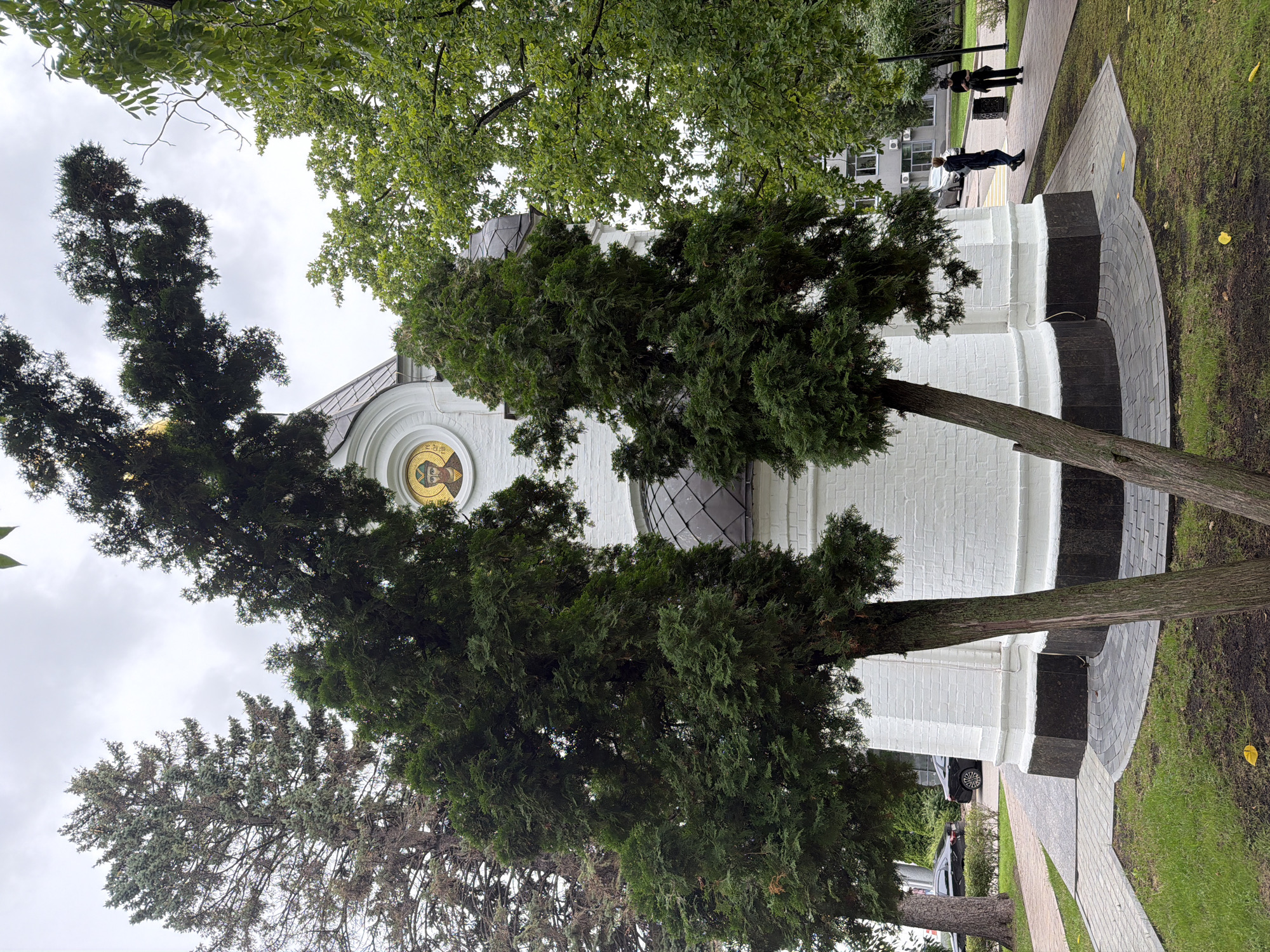 The image depicts an outdoor scene featuring a white brick building, possibly a chapel or part of a religious institution, with a domed roof covered in dark gray shingles. Two trees with dense green foliage are prominently positioned in the foreground, partially obscuring the building. A circular mosaic icon of a male figure is visible on the side of the building. The building is surrounded by a paved walkway and a grassy area. In the background, several people are walking on the path, and two cars are parked on a road to the left of the scene. The environment appears to be a park or a public space within a city, characterized by overcast skies and lush greenery. The location is identified as Nizhny Novgorod, Russia.