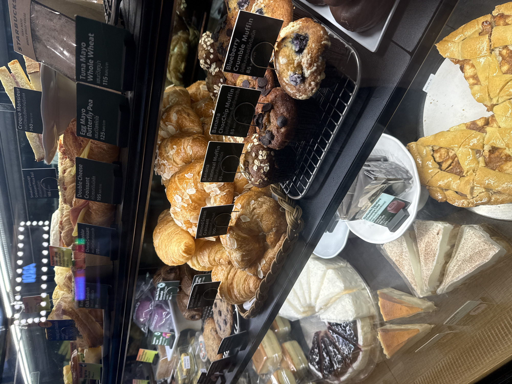 The image shows a display case filled with baked goods and desserts, likely in a cafe or bakery. The items include croissants, muffins, cookies, cakes, and a large apple tart. Labels for some of the items are visible, with prices indicated in Thai Baht. The environment appears to be indoors with artificial lighting illuminating the products. Some of the labels suggest that the location is in Thailand, specifically mentioning "Nong Prue Subdistrict, Thailand" in one of the cropped hints, which is a subdistrict of Pattaya City. There are no people visible in the image. The display case is organized and clean, showcasing a variety of food items.
