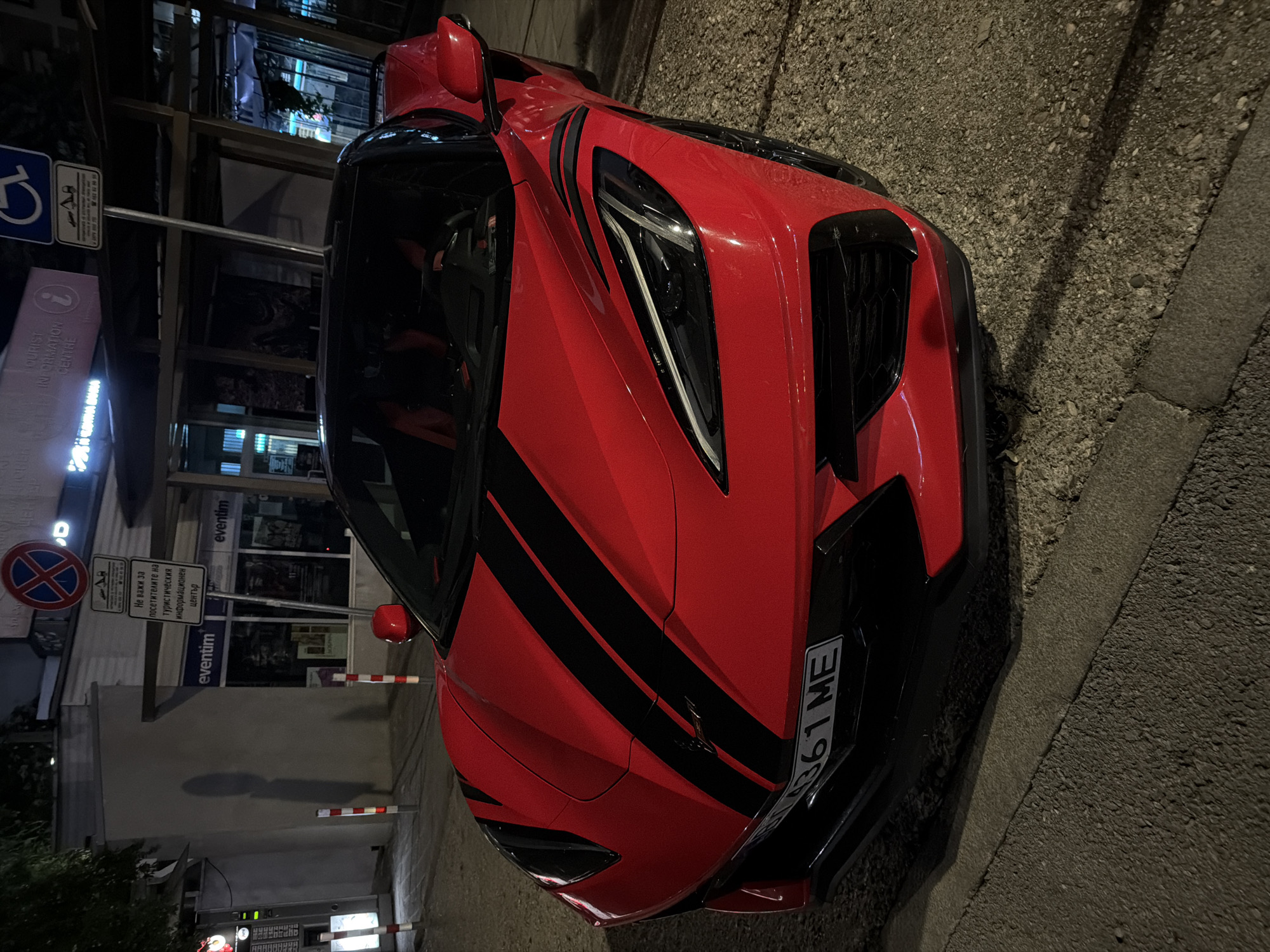 This image captures a red Chevrolet Corvette parked on a street at night in Veliko Tarnovo, Bulgaria. The car features black racing stripes on its hood and is angled towards the viewer, showcasing its front end. In the background, a building with "eventim" signage is visible, along with various street signs. One sign, likely a parking prohibition, displays a red circle with a diagonal cross, and another indicates "Tourist Information Centre." The street surface appears to be a mix of asphalt and gravel. No people are visible in the frame, and no actions or interactions are taking place. The overall scene depicts a nighttime urban setting in Bulgaria with a prominent sports car as the main subject.