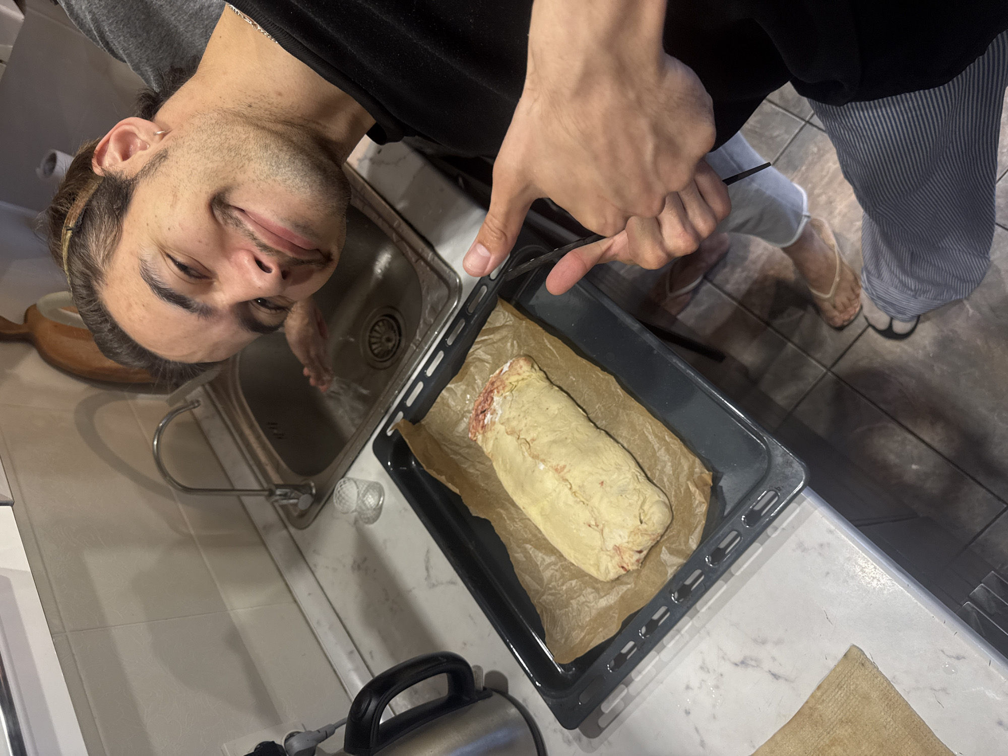 A man is standing in a kitchen, taking a picture of a meatloaf on a baking sheet. He is smiling at the camera. He is wearing a black shirt and striped pants. The kitchen has white tile floors and white countertops. A sink is visible to the left of the baking sheet.