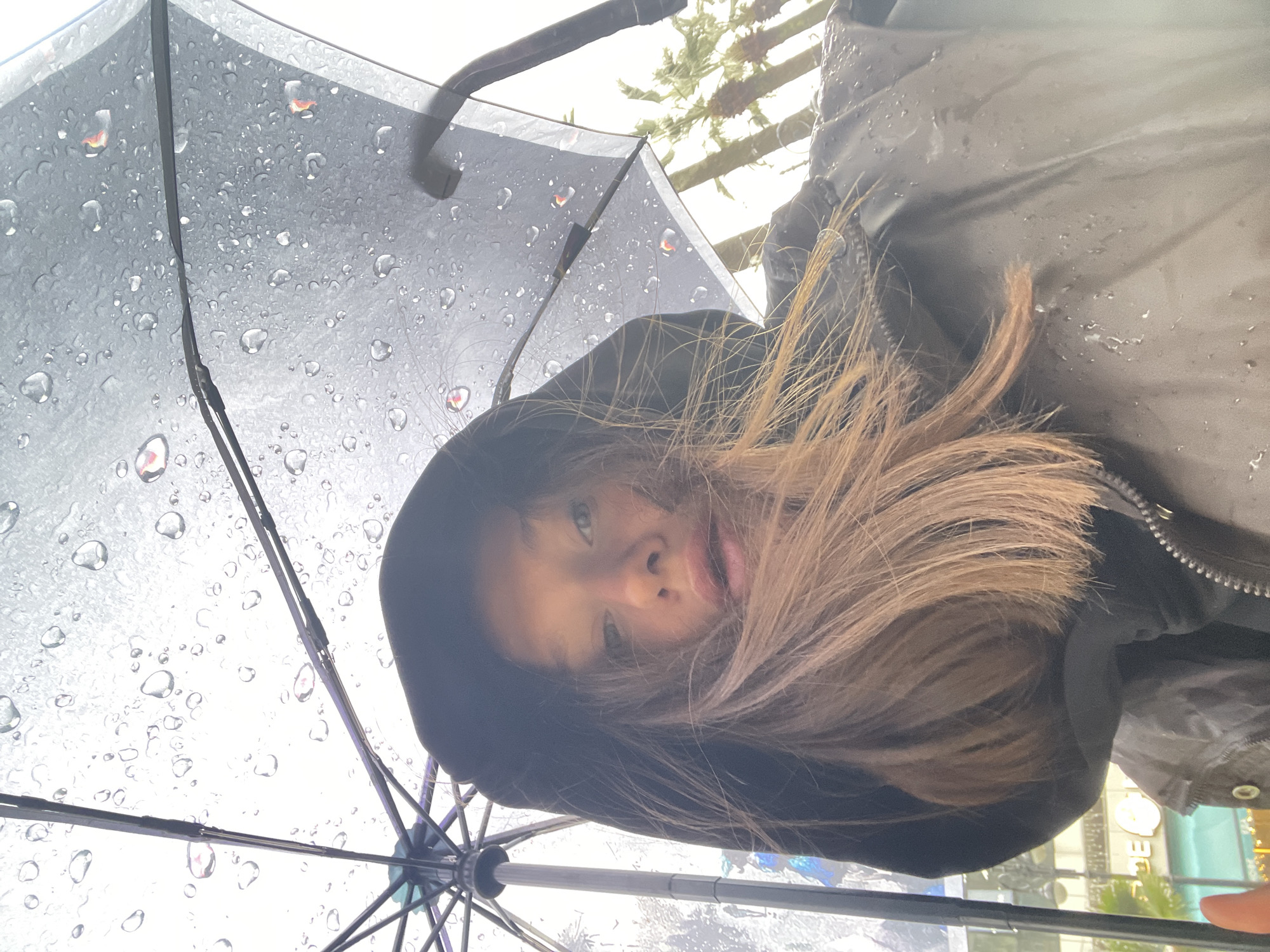 A woman is standing under an open black umbrella on a rainy day. The umbrella's surface is covered in raindrops. The woman has long, light brown hair and is wearing a black hooded jacket. The photo appears to be taken from a low angle, looking upwards. The background shows blurred buildings and trees, suggesting an urban environment. Based on the reflection in the water droplets on the umbrella and other details in the image, the location is identified as the city of Sochi, Russia.