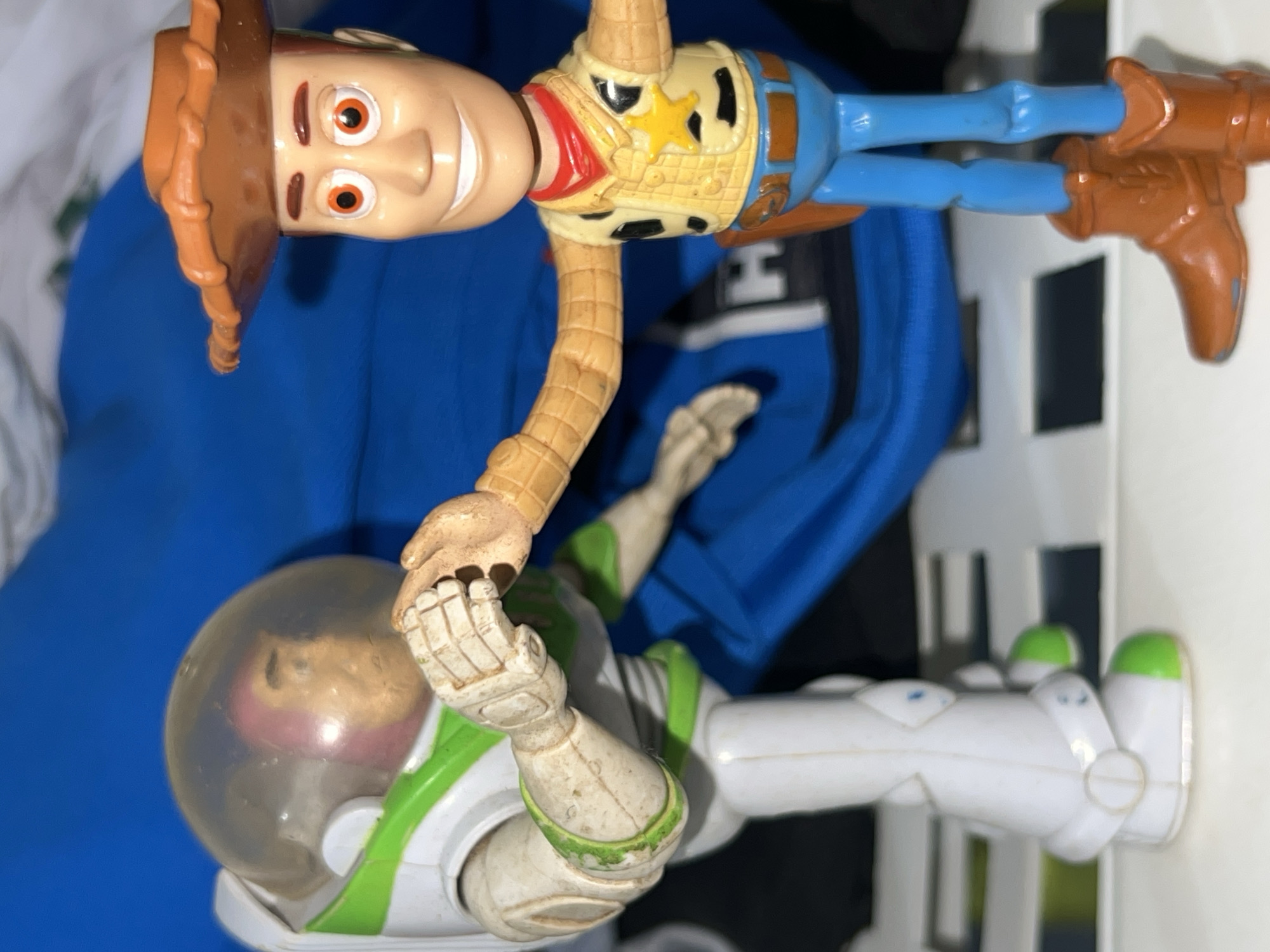 An image captures two iconic toy characters from Disney Pixar's Toy Story: Woody and Buzz Lightyear. The picture is oriented sideways, with Woody on the upper left and Buzz Lightyear on the lower right, seemingly interacting.
Woody, the cowboy doll, is positioned with his head, torso, and arms clearly visible. He wears his signature brown cowboy hat, has a light-colored face with orange eyes and a wide, smiling mouth. His outfit includes a red bandana, a yellow vest with black cow spots and a golden sheriff's star, and blue pants. His arms, made of a flexible, rope-like material, extend towards Buzz.
Buzz Lightyear, the space ranger action figure, is shown below Woody, appearing to be held by Woody's hand. He is dressed in his white and green space suit, complete with a clear dome helmet. His face is visible through the helmet. Buzz's suit, particularly his hands and forearms, shows signs of wear, scuffs, and dirt, indicating prior use.
The toys are set against a soft blue fabric background on the left, possibly a blanket or clothing item. On the right, a white structure with dark vertical elements, possibly part of a shelf or toy storage, is visible. The lighting appears to be bright and direct, suggesting an indoor setting. There is no visible text in the image.