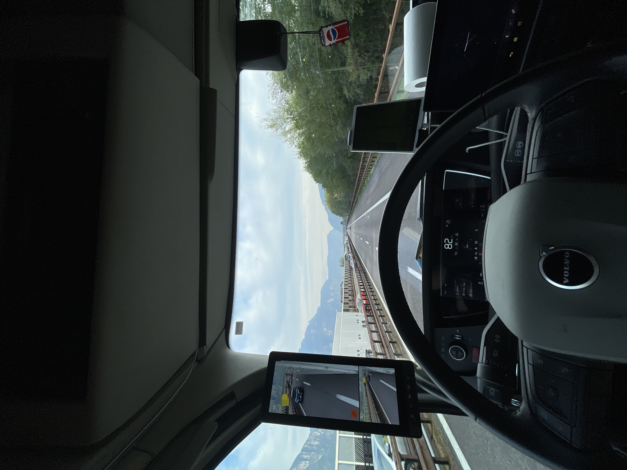 The image depicts the interior of a Volvo truck cab, providing a view of the road ahead through the windshield. The highway is bordered by barriers, and beyond the road, mountains can be seen. The speedometer indicates a speed of 82 km/h. A Pepsi-themed air freshener is suspended from the windshield. A screen displays a view from a side-view camera. The photo was likely taken in Nave San Rocco, Italy, based on location information.