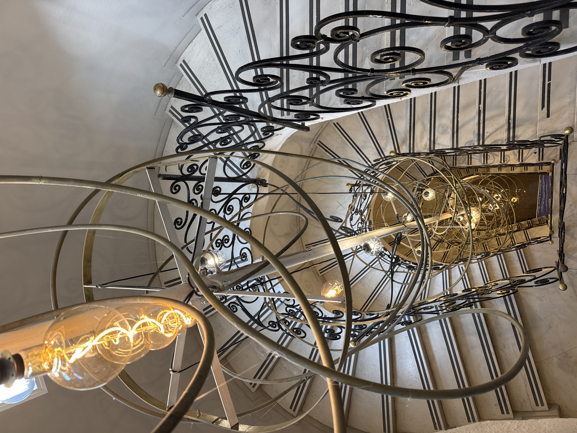 The media file presents an overhead, downward-looking view into a grand spiral staircase, likely within an elegant building in Isola delle Femmine, Italy. The central subject is an elaborate, multi-tiered light fixture suspended in the core of the staircase. This fixture is composed of numerous concentric and intersecting metal bands, appearing brass or gold-toned, housing multiple illuminated Edison-style filament bulbs and smaller clear glass bulbs that emit a warm, golden glow.
The staircase itself features light-colored treads, possibly marble, each adorned with distinctive parallel black strips, which could be anti-slip measures or decorative accents. A prominent black wrought iron railing, intricately designed with various scrollwork patterns, winds its way down the spiral. Small, spherical, gold-colored finials are visible on the railing posts.
The surrounding walls are a plain, light-textured surface, providing a neutral backdrop to the detailed ironwork and the glowing light fixture. No people are present, indicating a static architectural scene. The dramatic high-angle perspective emphasizes the spiraling form and the ornate details. A small, rectangular sign with dark background and light-colored, illegible text is visible attached to the lower part of the light fixture's frame or adjacent railing section. The time of day is not explicitly clear, but the illuminated bulbs suggest indoor lighting is primary, possibly during evening or in a dimly lit interior space.
