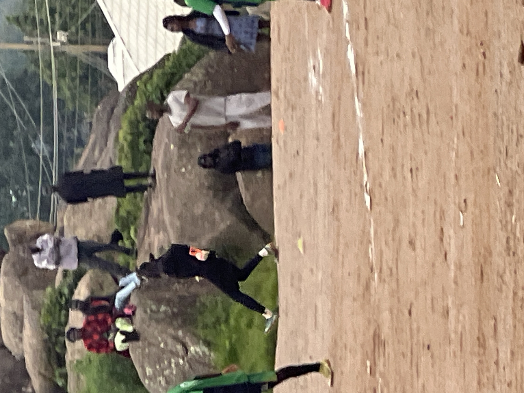 The image depicts a group of people gathered outdoors in what appears to be a dusty, open area, likely a sports field or a park, with a rocky, natural landscape in the background. The location is identified as Jos, Nigeria. In the foreground, a person in a black outfit is captured mid-stride, appearing to be playing a sport or engaged in an activity that involves movement. This individual is wearing dark pants and a dark top, with visible sneakers. To the left, another person is partially visible, wearing a green garment and dark pants.
In the middle ground, several individuals are positioned on and around a large rock formation. One person in a white shirt and light-colored pants stands facing away from the camera. To their right, a woman in a blue patterned dress and a dark jacket is also visible. Behind them and further to the left, two people are seated on the rocks; one wears a red and black patterned shirt, and the other is in a dark outfit. Further back on the rocks, another individual in a black robe is standing. In the distance, trees and power lines are visible, suggesting an urban or semi-urban environment. The ground is predominantly brown and appears to be dirt or sand, with some white markings that could indicate a sports field boundary. Small yellow and orange cones are scattered on the ground, further suggesting a sporting event or training session. The overall lighting suggests it is daytime.