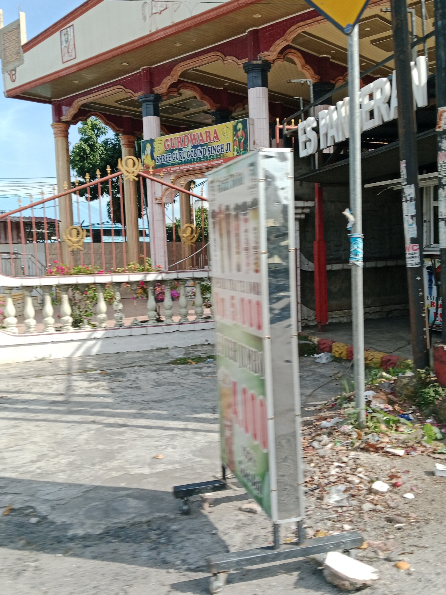 This image depicts a street scene in the City of Binjai, Indonesia, with a prominent building that appears to be a Gurdwara, a place of worship for Sikhs. The building has a red and cream facade with decorative arches and columns. A yellow sign with an arrow is visible at the top of the frame. In front of the Gurdwara, a metal fence with gold Sikh Khanda symbols is present. A large, vertical advertisement board is positioned in the foreground, on wheels, obstructing a clear view of the street and the building behind it. The advertisement board has a reflective surface and appears to display images or text. The ground in front of the building is paved with concrete and shows signs of litter and debris, including plastic bags, paper, and broken pieces of material. A tree is visible behind the fence. To the right of the Gurdwara, a sign with large white letters spelling "ES PAMPERAN" is mounted. The overall environment suggests a public area with commercial activity and potential neglect due to the presence of litter. The sky is partly cloudy.