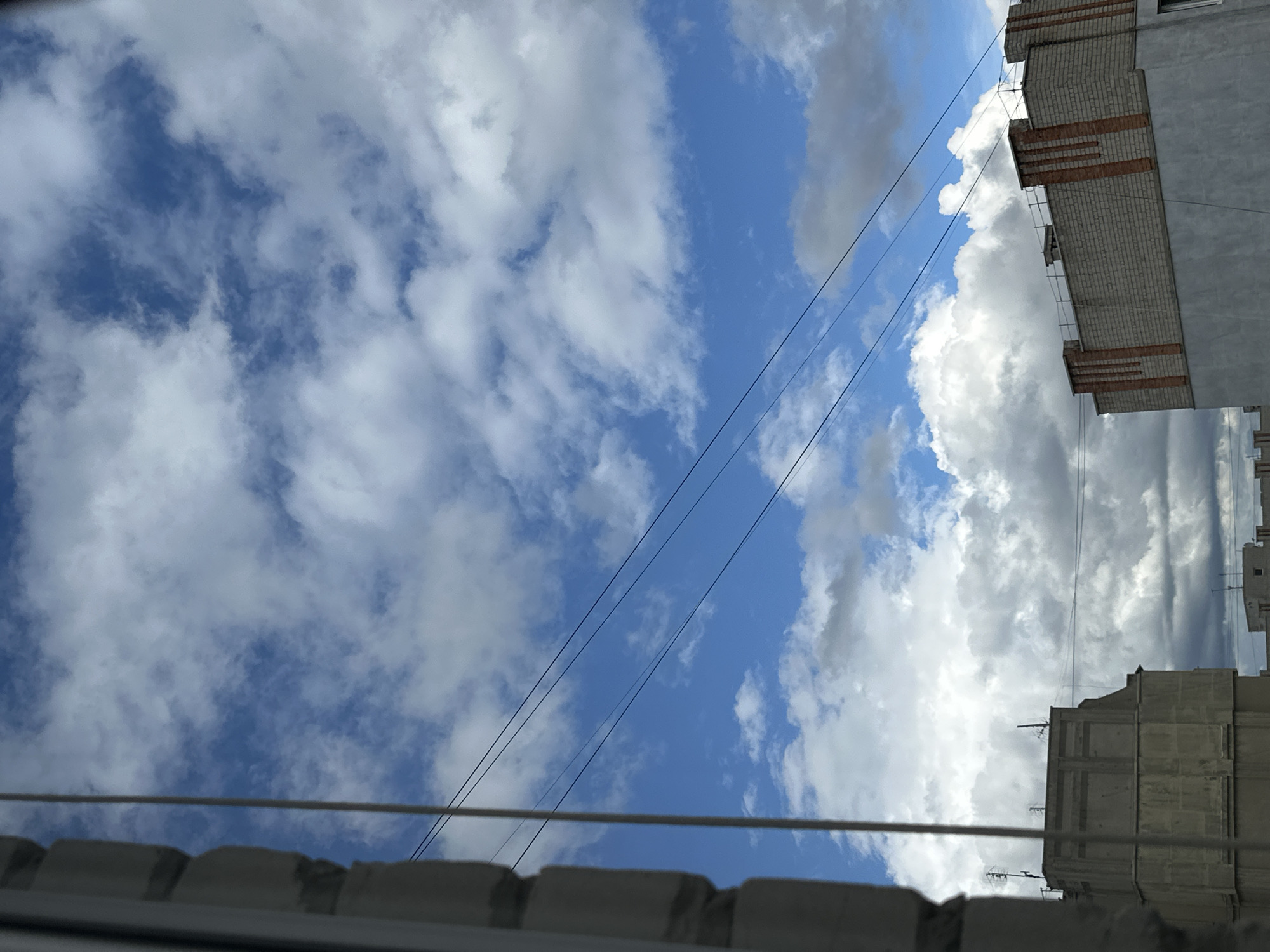 The image shows a view looking upwards towards a partly cloudy sky with blue patches, with the presence of buildings and utility lines. The buildings are located in Unknown, Unknown. Two buildings are visible, one constructed of grey brick with horizontally oriented red brick accents, and the other is built of grey concrete blocks. Both buildings are multistory and suggest an urban or suburban environment. Utility lines stretch across the sky, and a portion of a building rooftop is partially visible in the foreground.
