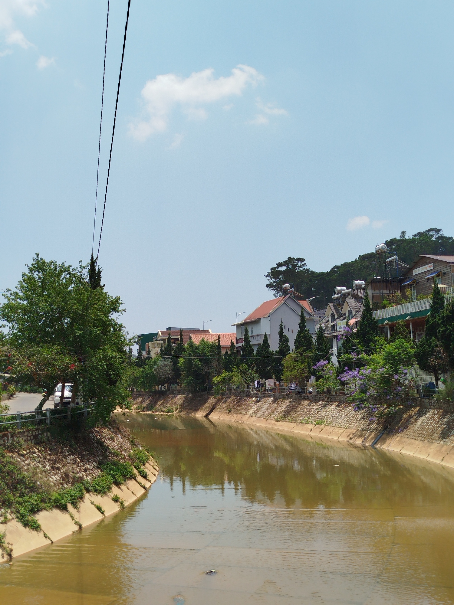 This image captures a canal running through Cam Ly - Đà Lạt, Vietnam. The water in the canal is a murky brown. Concrete and stone embankments line both sides. Lush greenery and trees grow along the banks and beyond, with residential buildings perched on the hillside in the background. A few solar water heaters are visible on rooftops. Power lines are noticeable in the upper left corner of the frame. The sky is blue with a few scattered clouds.