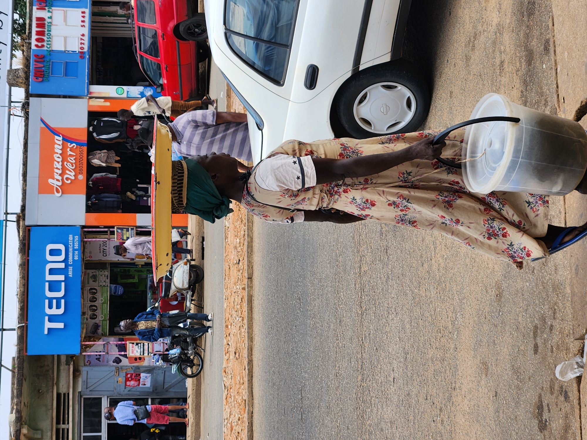 The image captures a street scene in the city of Unknown, Unknown. An elderly woman is the main subject, she is dressed in a floral print dress and carrying a clear plastic bucket. She has a headscarf wrapped around her head. The woman appears to be walking along the edge of the road, near a curb or sidewalk. The road itself takes up a significant portion of the frame.
In the background, storefronts are visible on the left side of the image, with several commercial establishments identified by signage. One is a shop called "Arizona Wears", and another advertises "TECNO" Arise Communication. A red vehicle and a white car are partially visible in the scene, parked next to the buildings. There are people standing and sitting near the shops. A motorcycle is parked on the side of the road.