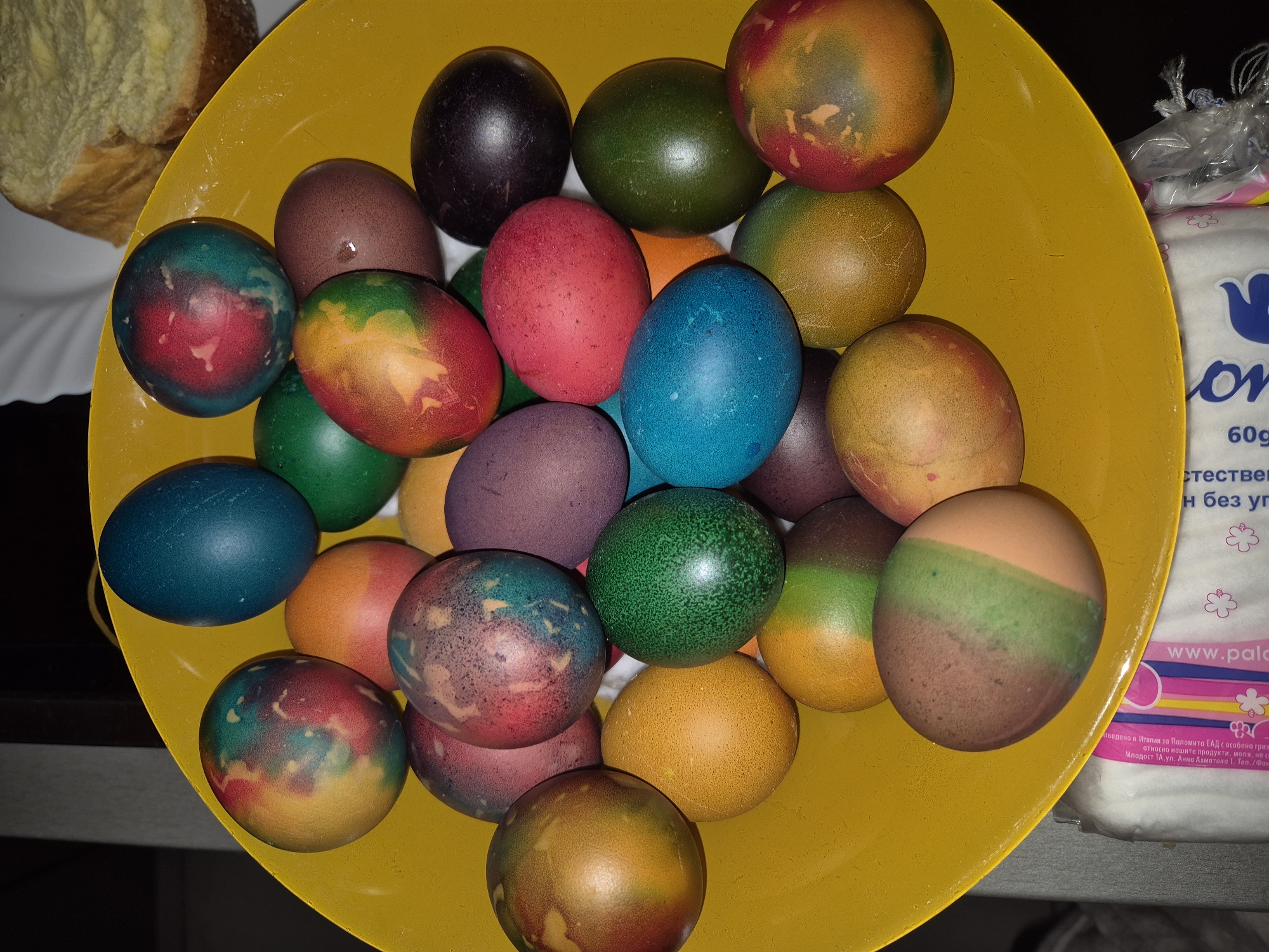 A top-down, close-up shot displays a yellow bowl filled with a variety of dyed Easter eggs in various colors, including blue, green, red, purple, and orange, some with mixed colors, suggesting a traditional egg-dyeing process. To the left of the bowl, a piece of bread-like pastry sits on a white plate. Adjacent to the bowl on the right, there's a package with text, which includes "60g" and "www.palo," indicating a product and a website address. The text in Cyrillic suggests a location in Bulgaria, possibly Pavlikeni, given the mention of Паломита ЕАД (Palomita EAD). The setting seems to be an indoor location, likely a home or kitchen.