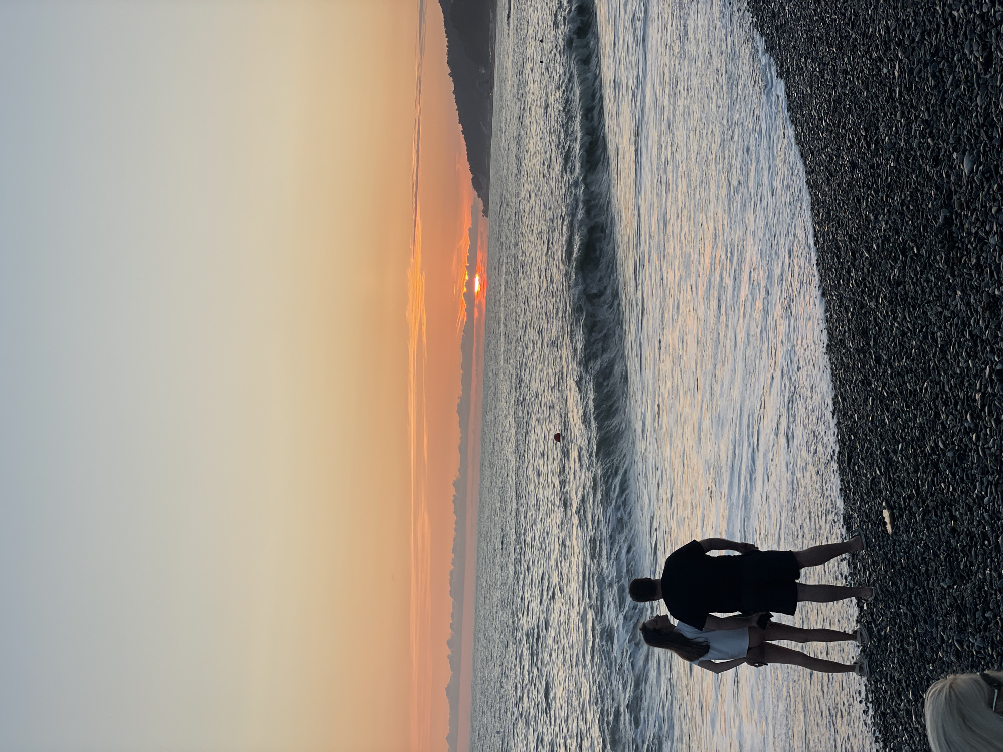 A man and a woman stand on a pebble beach on the Black Sea coast in Sochi, Russia, facing the ocean at sunset. The sky is a gradient of soft blues and oranges, with the sun partially obscured by clouds near the horizon. Gentle waves lap at the shore, and a few buoys are visible in the water. The man is wearing a black t-shirt and shorts, while the woman is in a white t-shirt and shorts. Their backs are to the camera as they gaze out at the sea. The beach is covered in dark, smooth pebbles. In the foreground, a person's head and shoulders are visible, also facing the sea. The overall scene evokes a sense of calm and tranquility.