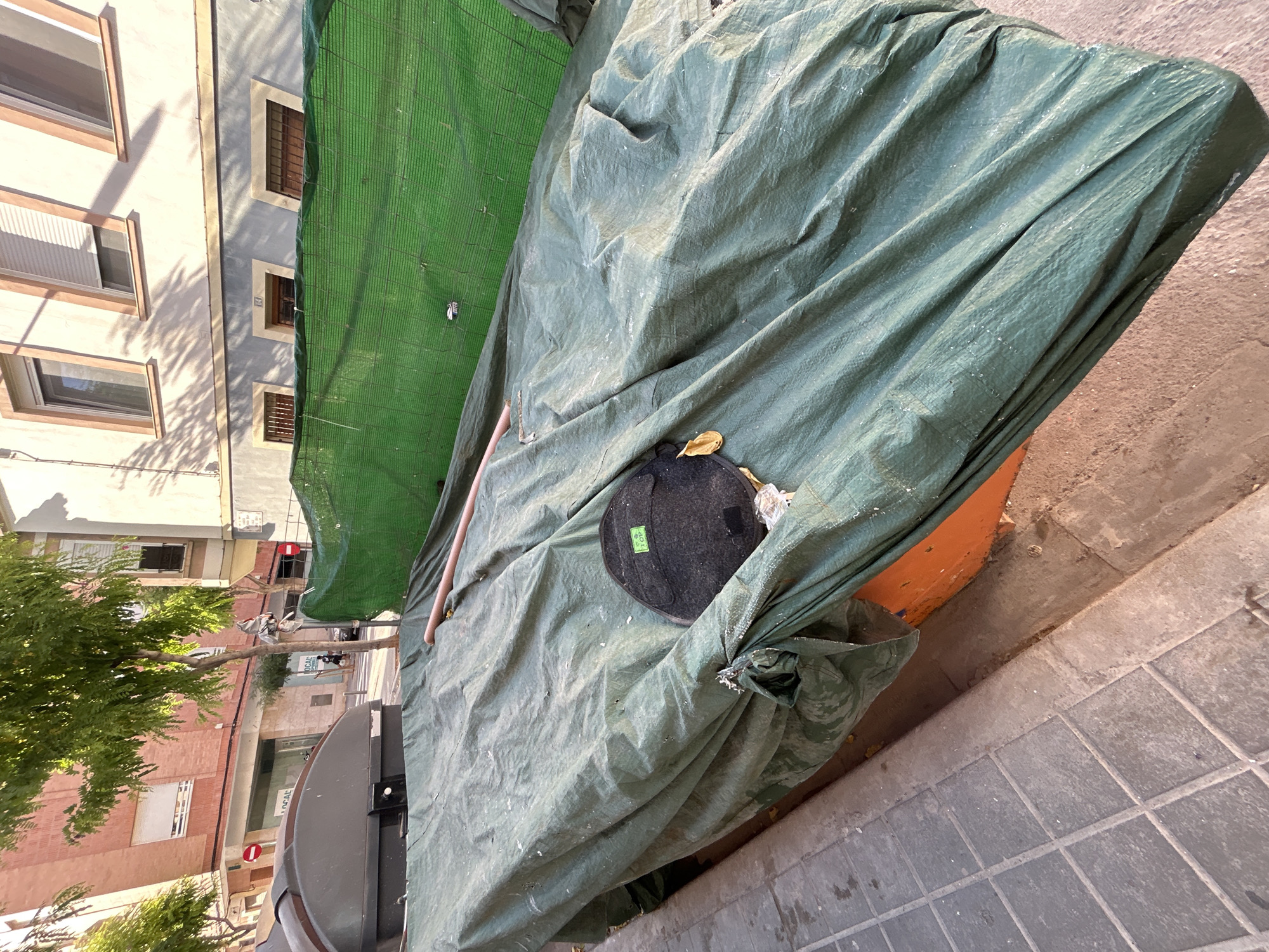 This is an eye-level, full shot of a street scene in Castelló De La Plana, Spain, during the day. The image is dominated by a large, green tarp covering what appears to be an outdoor object or structure. The tarp is heavily wrinkled and draped, suggesting it is being used to protect something from the elements or for storage. A thin, light-colored pole is visible poking out from under the tarp.
On the tarp, a circular, dark-colored object with a green tag that reads "TOP" is placed. Next to it, a small, dry, yellow leaf rests on the green material. The tarp itself is draped over an orange-colored surface, hinting at the object it is covering.
To the left of the tarp, a portion of a building is visible. It has a light-colored facade with several windows, some of which have wooden shutters. Sunlight casts distinct shadows of tree branches onto the building's walls, indicating it is a sunny day. Further to the left, the leaves of a lush green tree are visible.
The ground in the foreground is paved with square, grey tiles. The scene appears to be on a sidewalk or street. The overall impression is one of an urban outdoor space, possibly undergoing some form of construction or renovation, or simply storing items. The lighting suggests it is midday. There are no people visible in the frame.