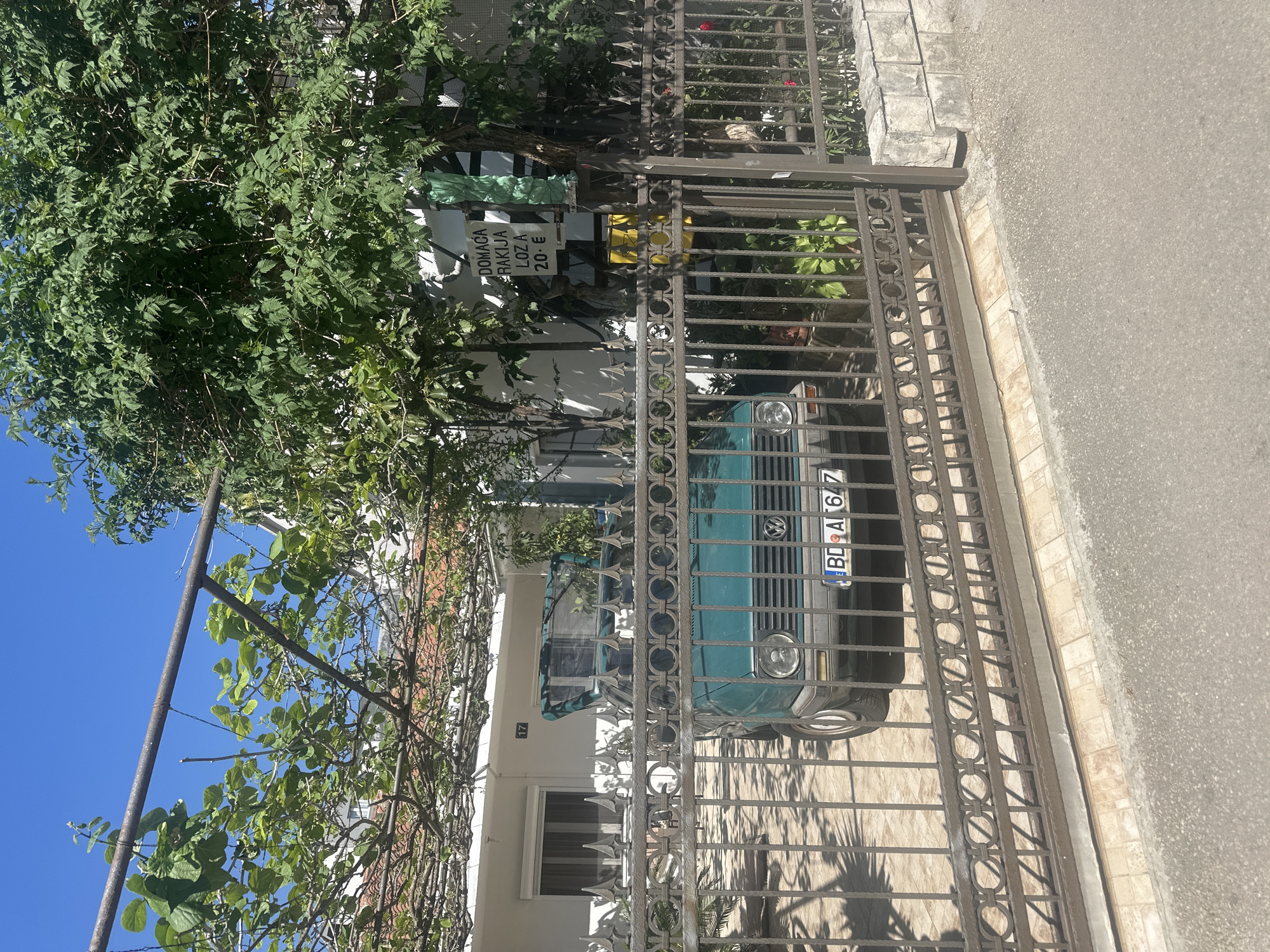 An outdoor, daytime shot of a property in Budva, Montenegro. A teal Volkswagen Golf is parked behind a dark metal gate, partially obscured by greenery. A white building with the number "17" visible above a window is behind the car. A sign that says "DOMACA RAKIJA LOZA 20 €" is attached to the gate, indicating homemade brandy is for sale. The sky is bright blue. The street pavement is visible to the right of the gate.